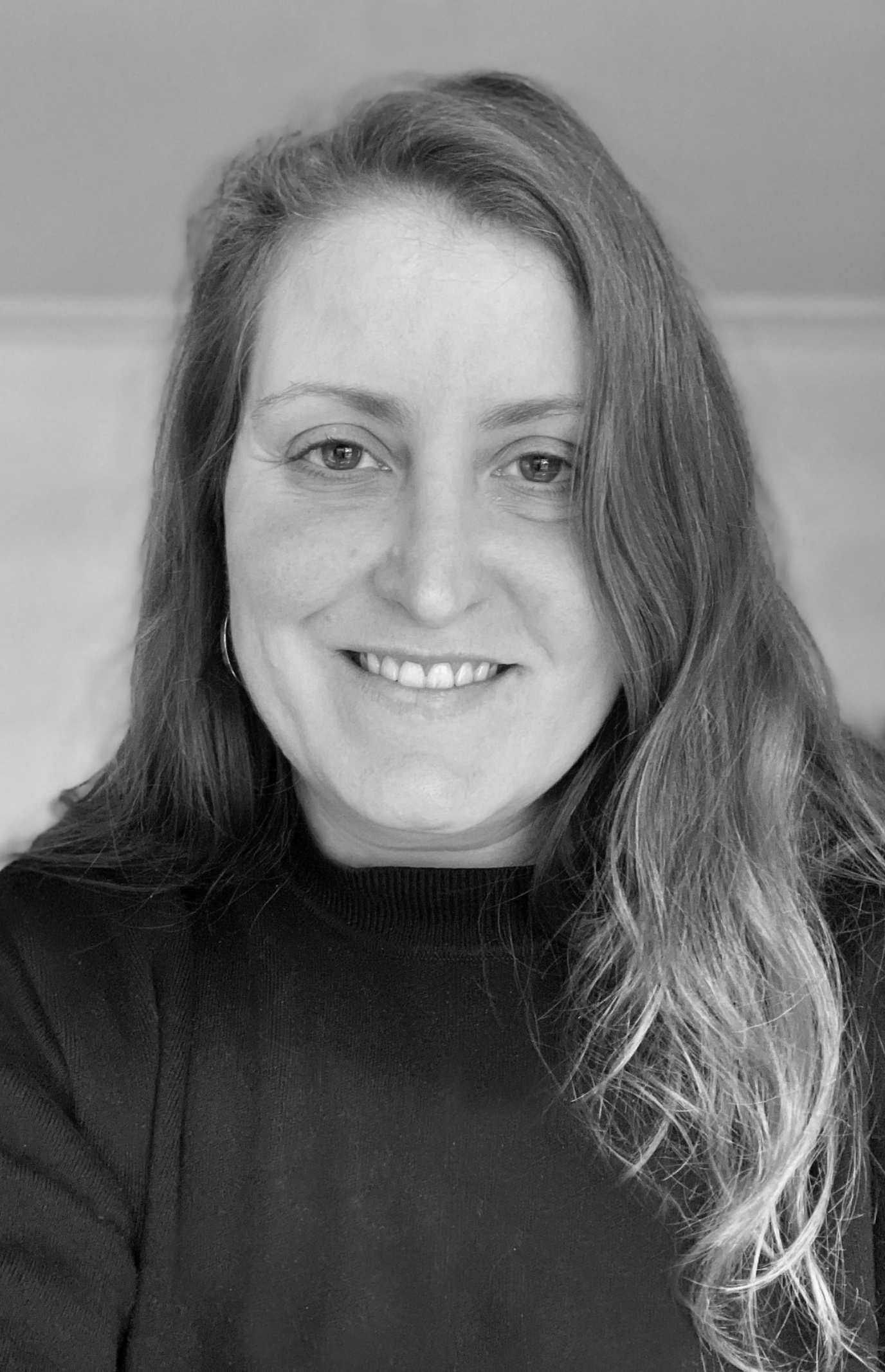 A close-up portrait of a woman with long wavy hair, smiling, wearing a black top, in a grayscale photograph.