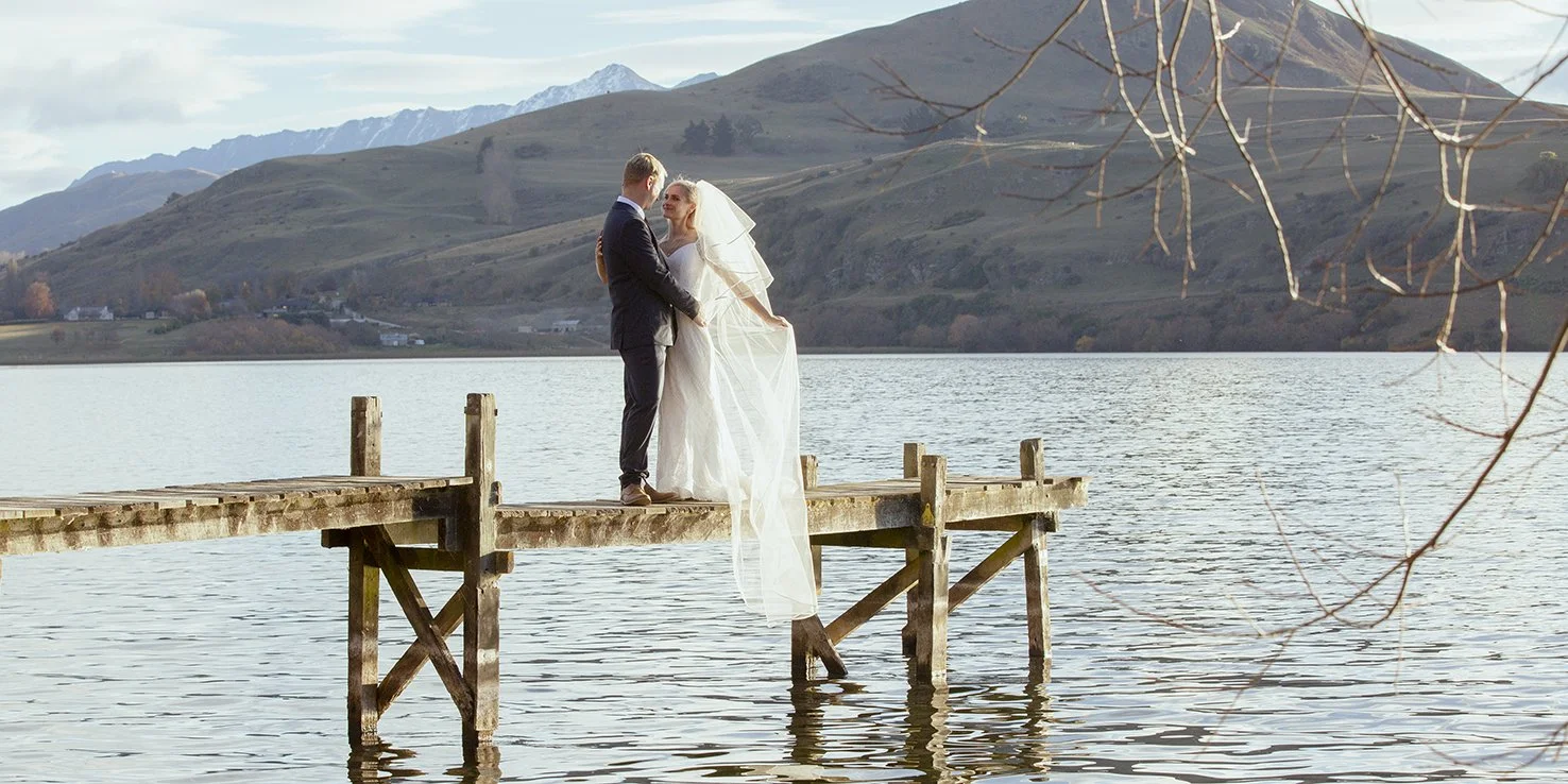 Newlyweds standing on a lakeside jetty with mountains behind them.