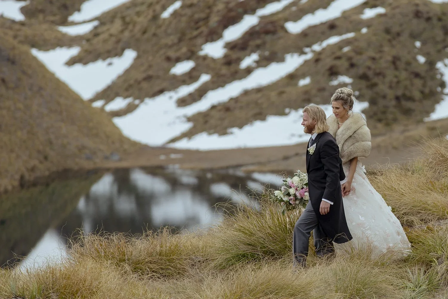 A newlywed couple walking beside an alpine tarn beneath snow-streaked mountains.