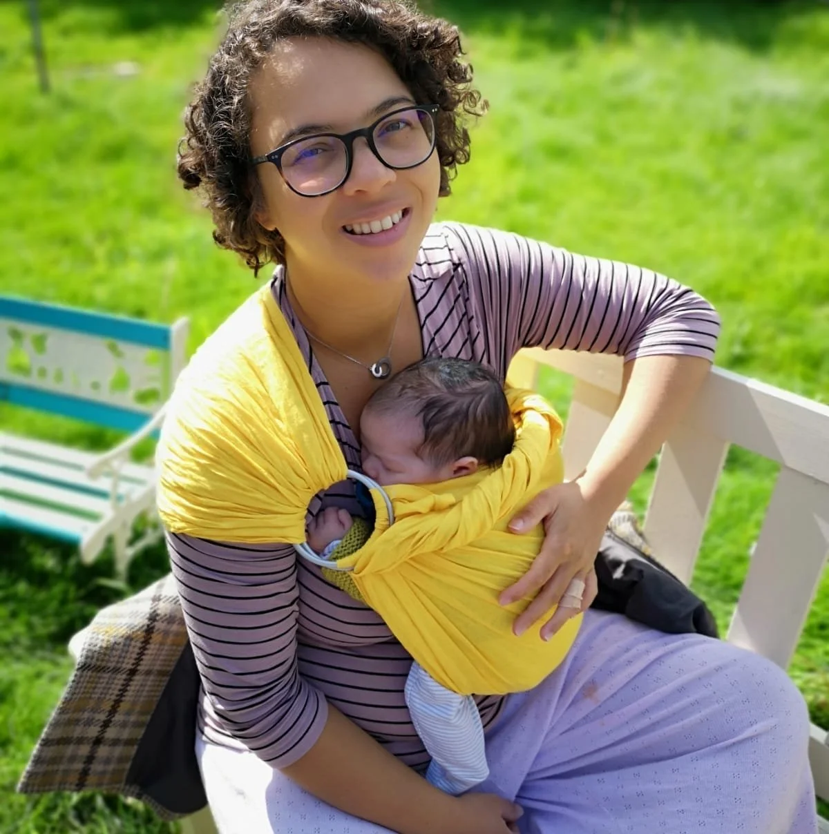 Ella sits on an outside bench smiling at the camera. She is wearing glasses and has short hair. She is wearing a bright yellow sling, which holds a newborn baby snuggled to her chest