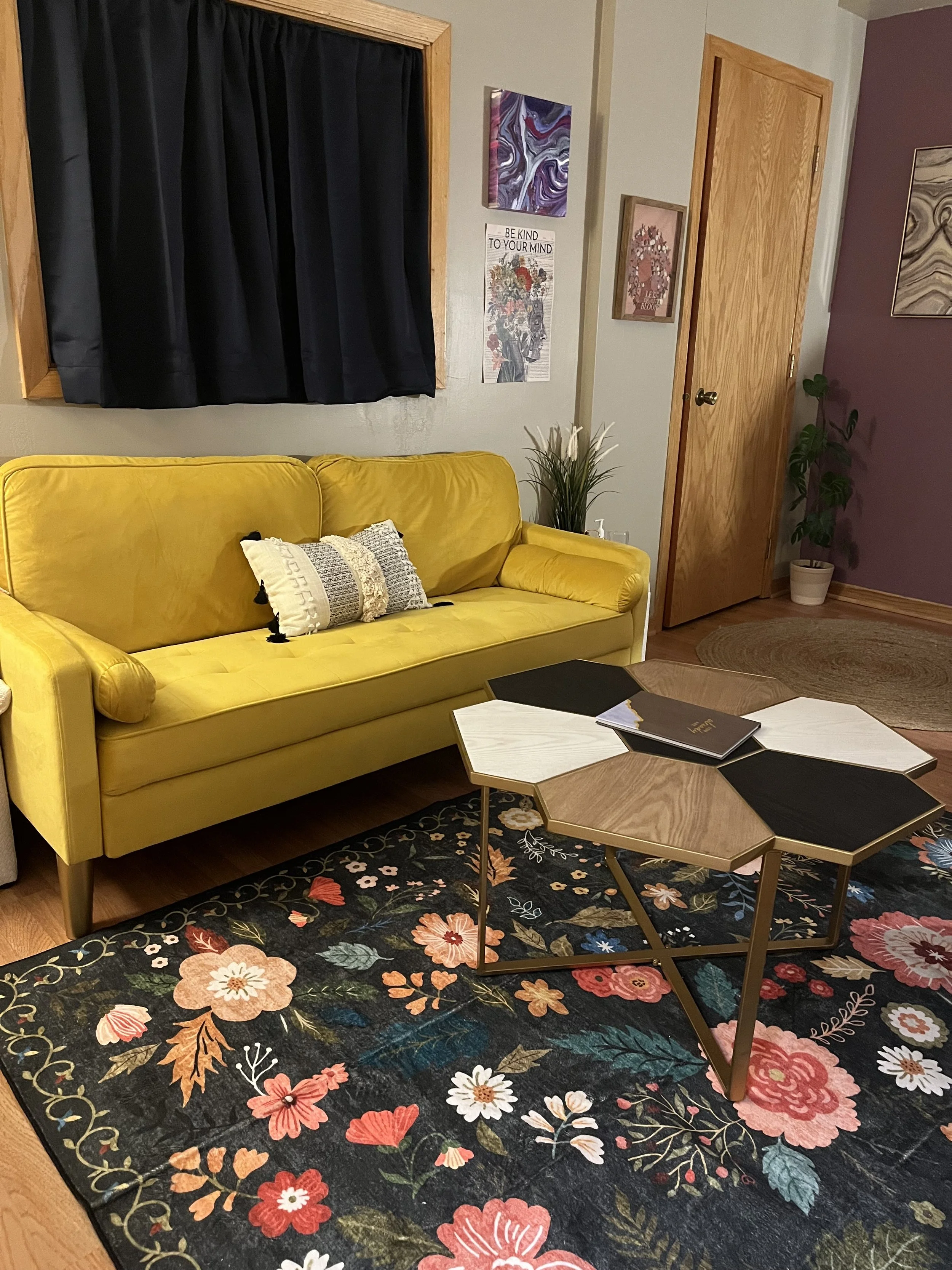 Living room with a yellow sofa, floral area rug, black and white coffee table, wall art, and a tall plant near a wooden door. Black curtains cover a window.