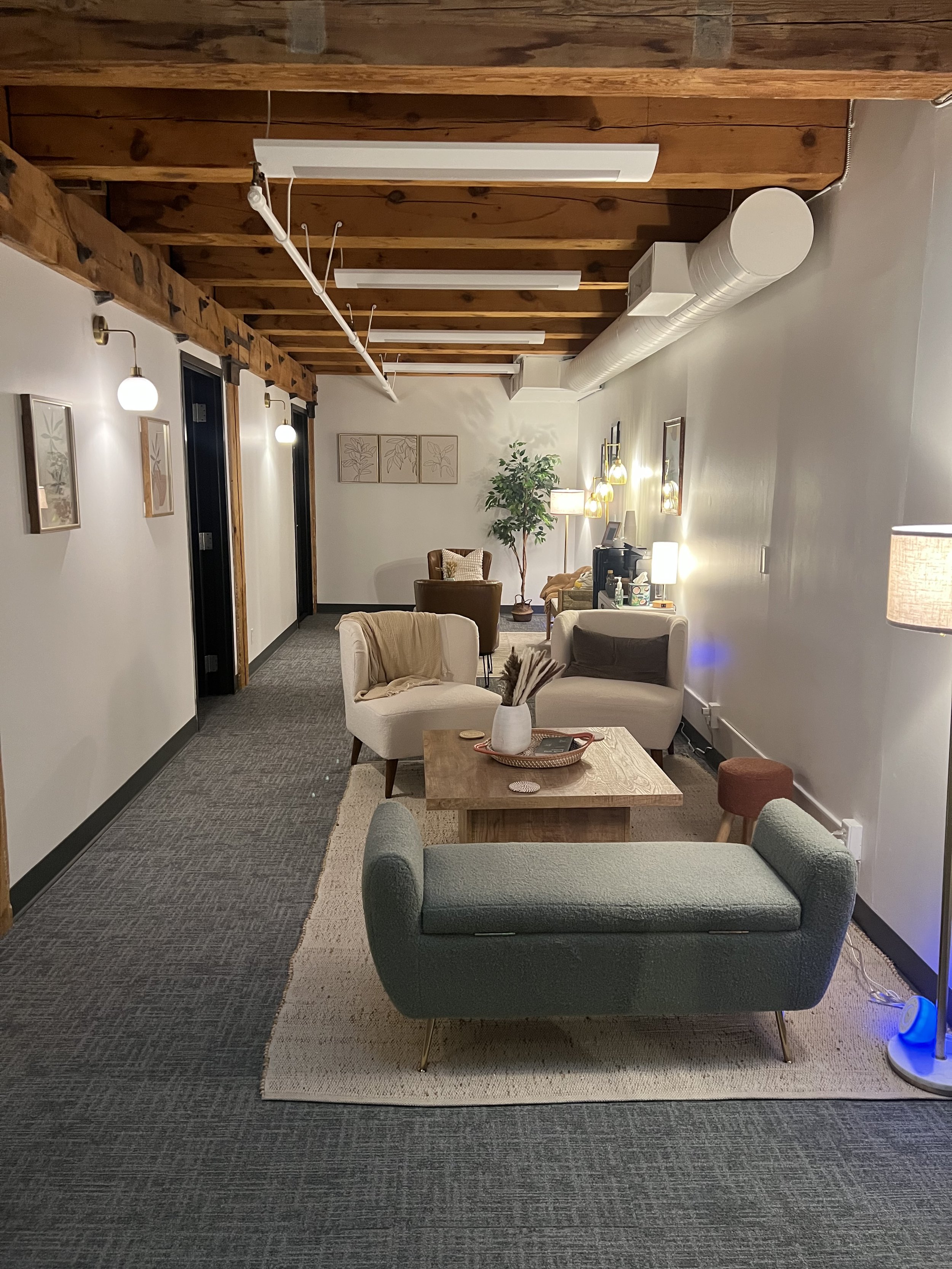A cozy living room with a window covered by white curtains, surrounded by string lights on a white brick wall. There is a gray sofa with pillows, a small round side table with snacks, a vintage armchair with a blanket, a white side table with decor and a clock, and shelves with pictures and decorations. The room has a warm, inviting atmosphere with various wall art and a rainbow pride flag.