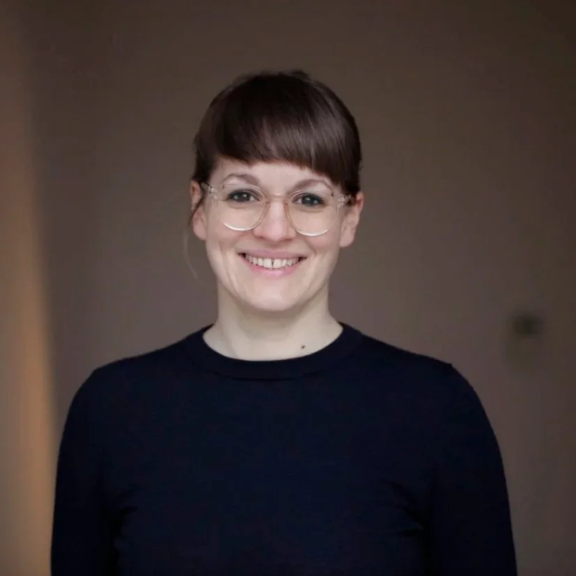 Frau mit Brille und braunen Haaren, lächelnd, vor neutralem Hintergrund.