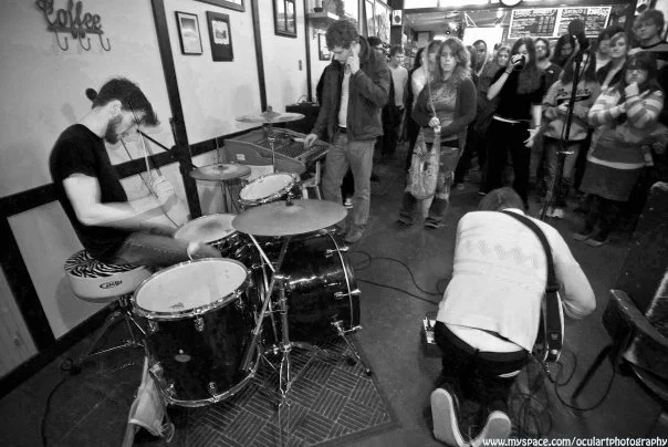 A drummer and guitarist perform live at a crowded indoor venue with an audience watching.
