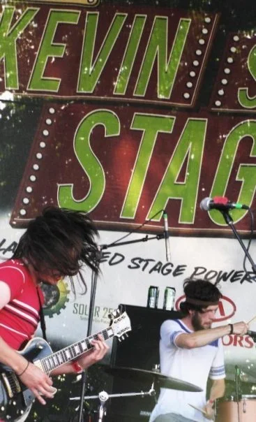Musicians performing on stage at the Kevin Stag event, with large green and red signage in the background.