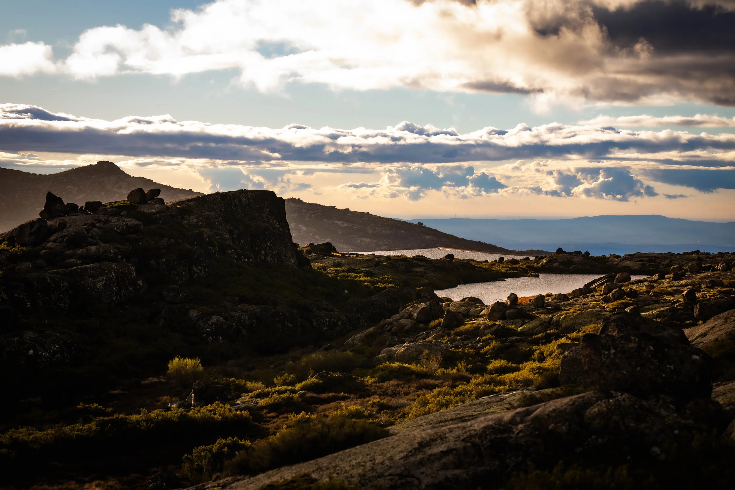 Een berglandschap met rotsen, een meer, en een bewolkte hemel tijdens zonsondergang.