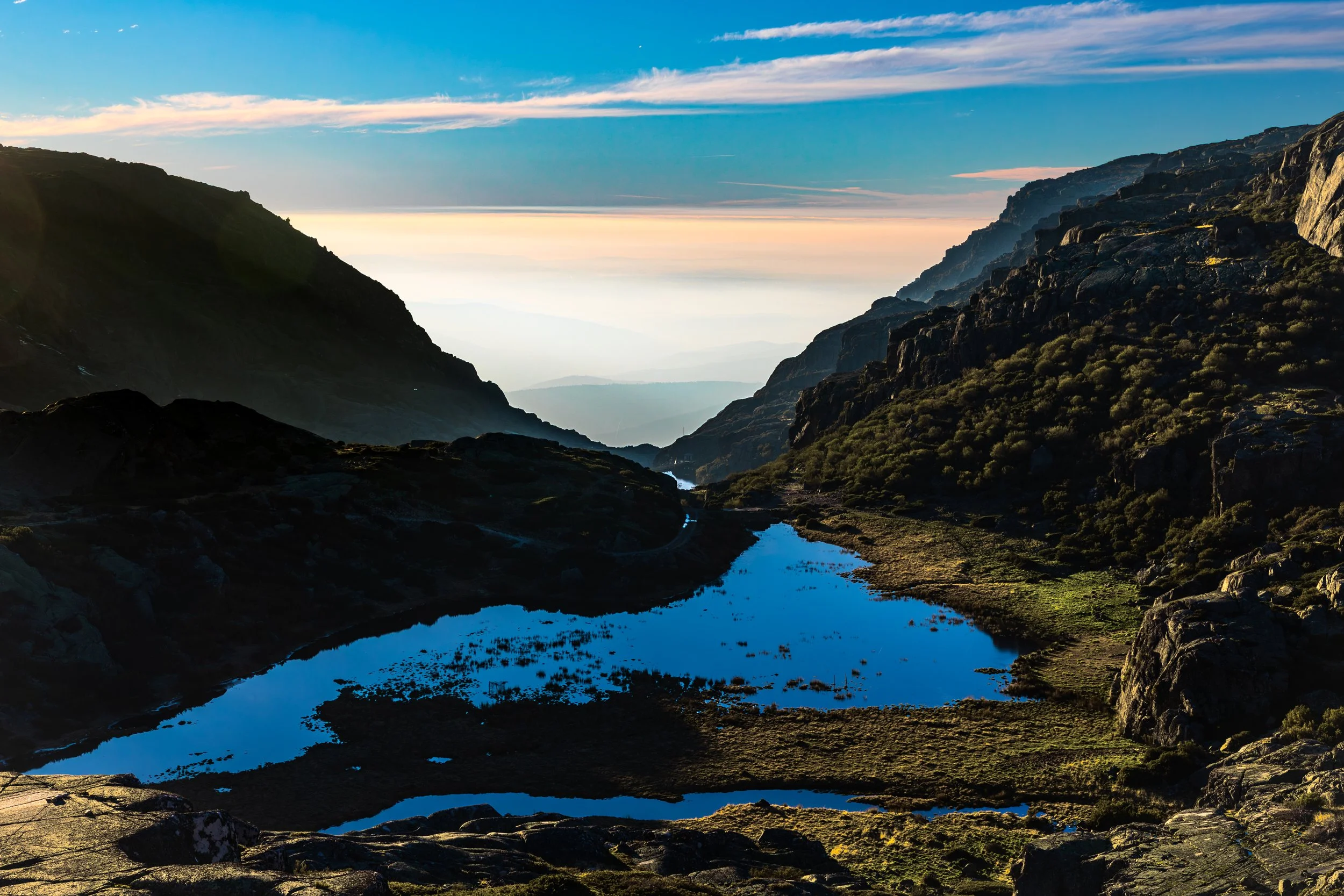 Een berglandschap met een kleine, heldere waterplas in de voorgrond, omlijst door rotsen en groene begroeiing, met bergen en een kleurrijke hemel op de achtergrond.