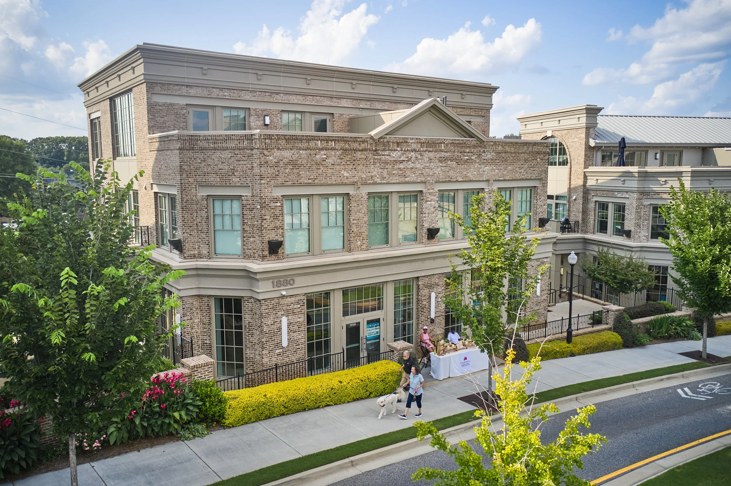 A multi-story brick residential building with greenery and sidewalk in front. People are walking their dogs and setting up a table outside. There are trees and street lamps along the sidewalk.