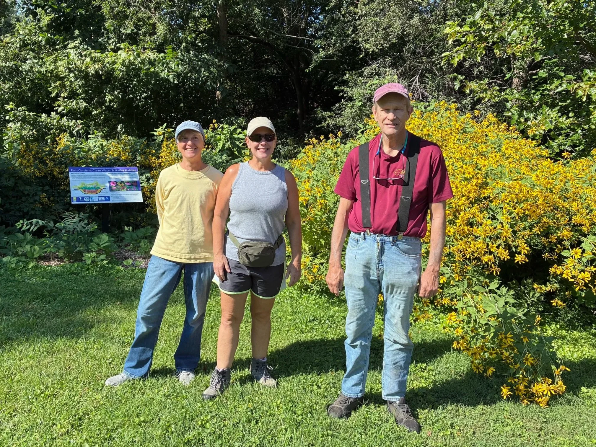 Three volunteers outdoors in the World Peace Gardens, two men and one woman, standing near gardening tools and plants, with trees and a residential area in the background.