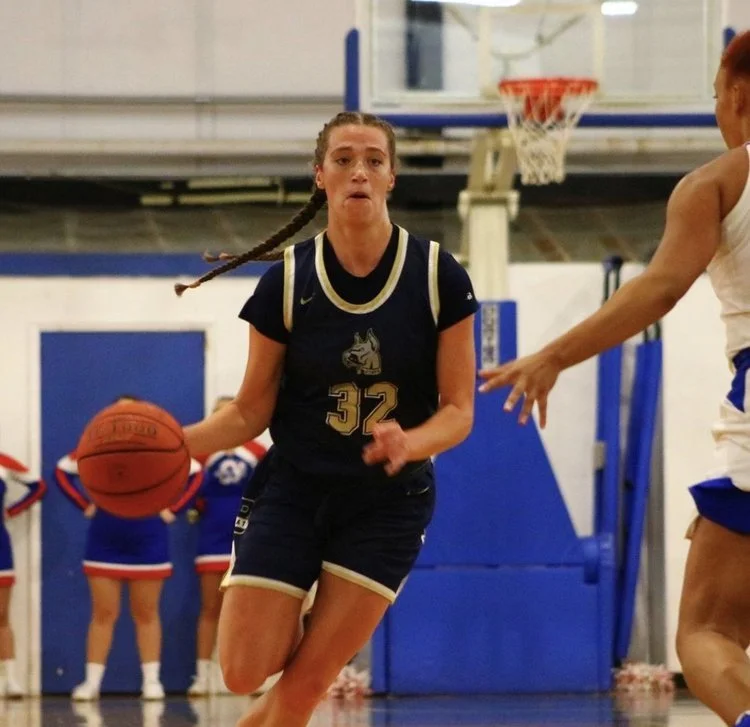 Female basketball player in a navy uniform dribbling the ball on a court with cheerleaders in the background.