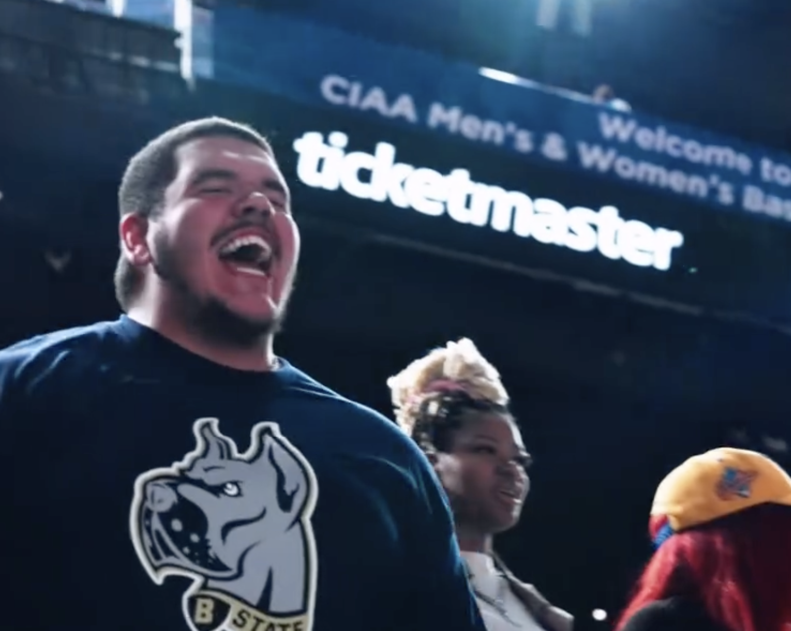 A group of people cheering at a sports event with a visible sign for Ticketmaster in the background. The man in the foreground is wearing a navy blue shirt with a dog mascot design and the text 'B State.'"