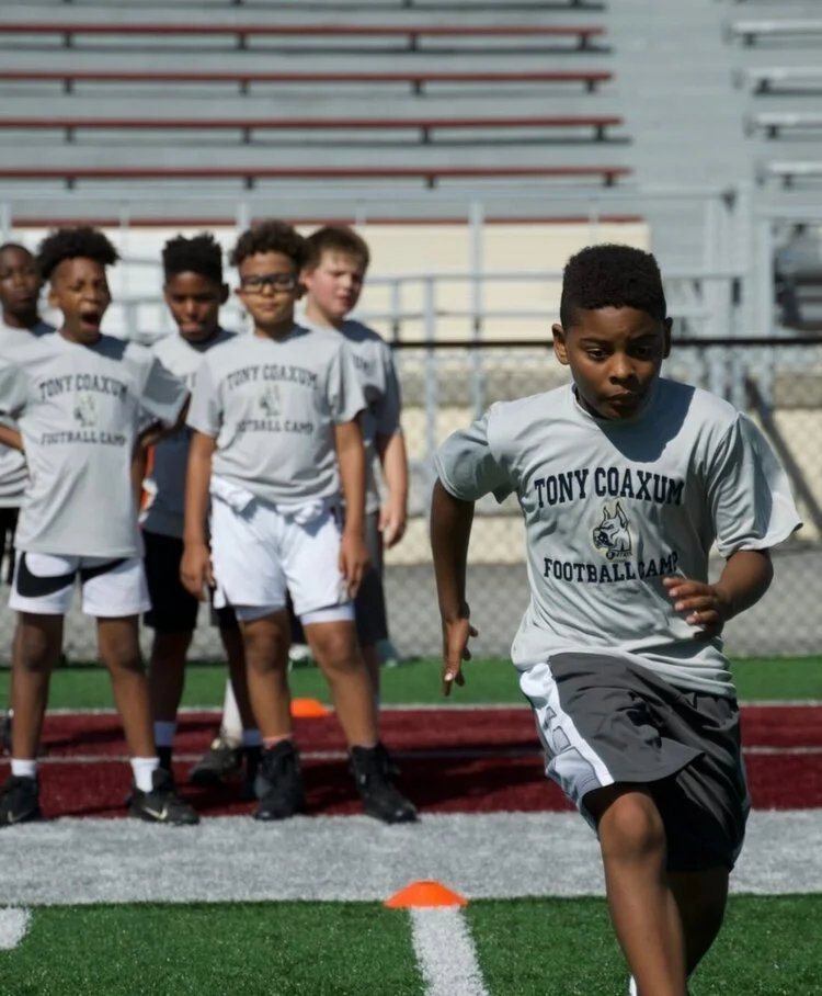 Kids participating in a football camp, wearing gray "Tony Coaxum Football Camp" t-shirts, one boy running in the foreground while others watch in the background.