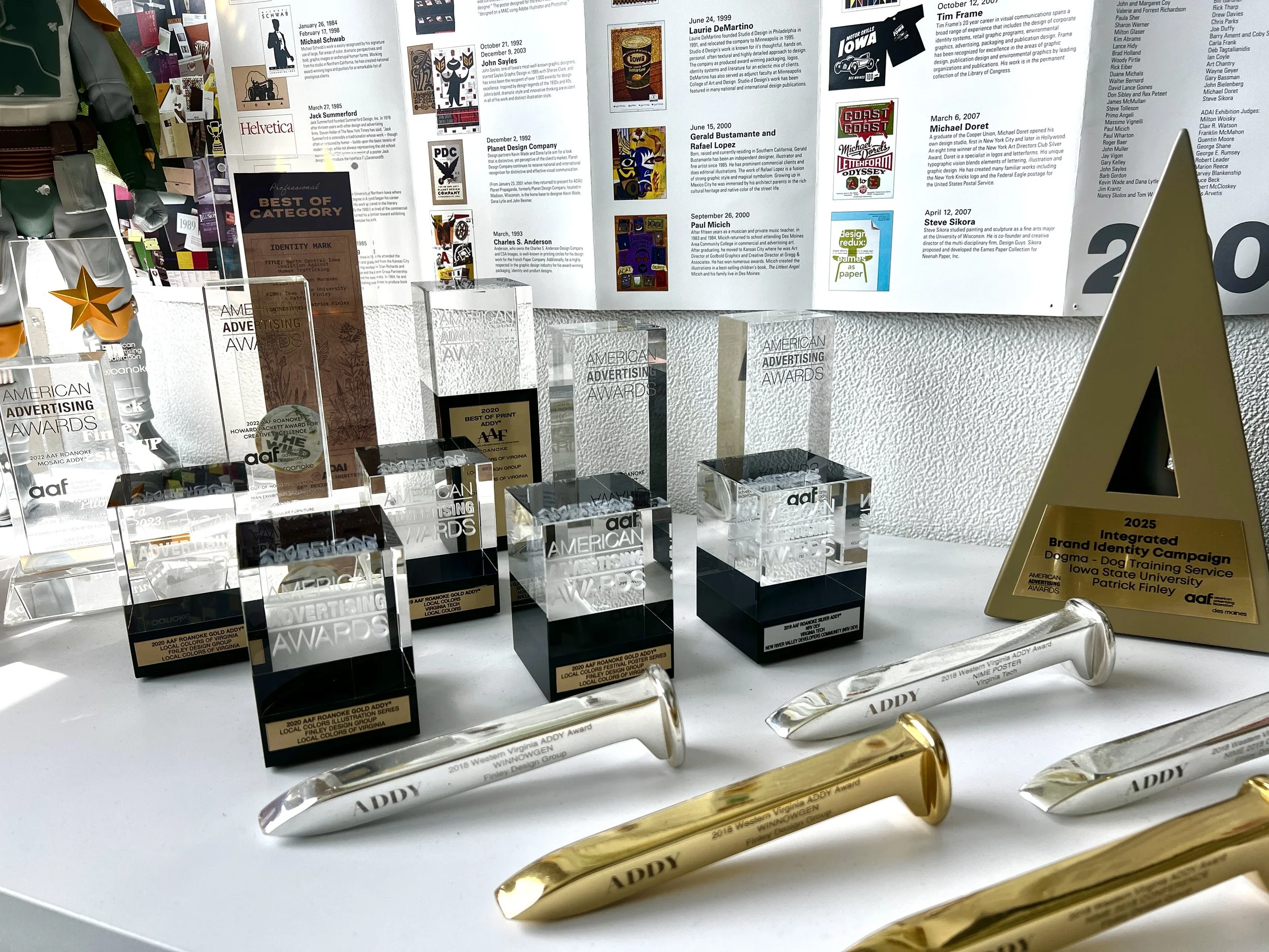 Awards and trophies displayed on a white table with a background of a bulletin board featuring various posters and notices, including winning categories and dates.