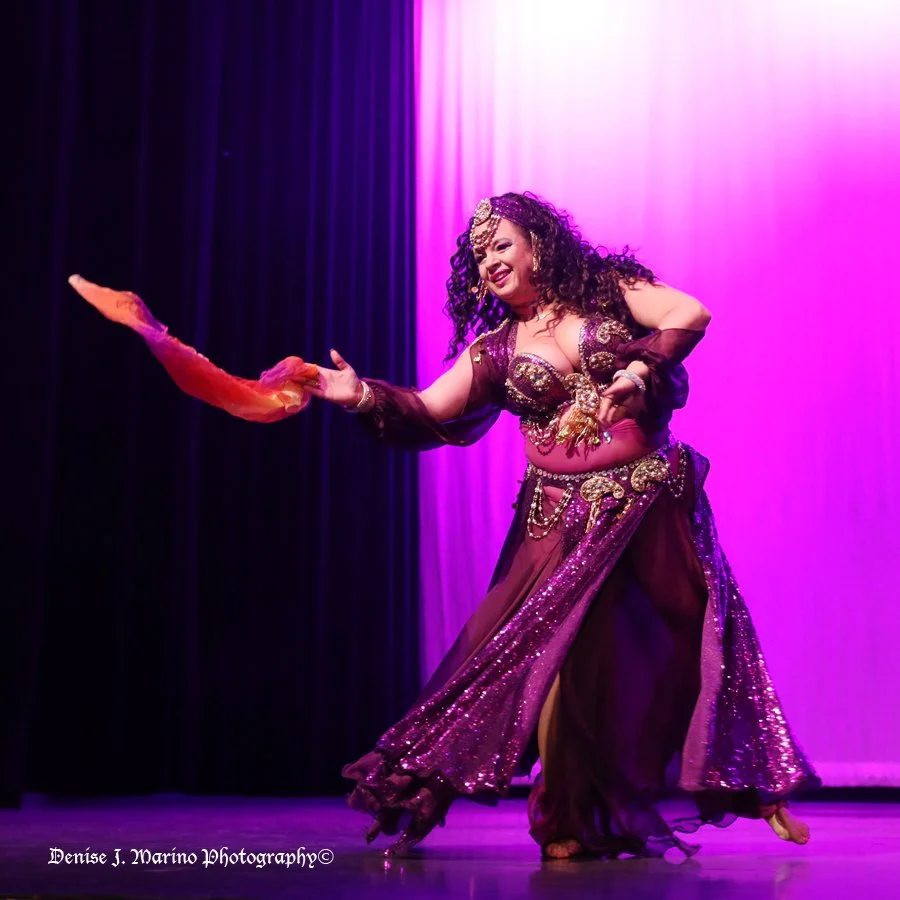 Two women dancing in a lively bar or restaurant, surrounded by seated patrons. One woman has gray hair with a red flower, wearing a blue dress with floral patterns. The other has long dark hair, wearing a red and white floral belly dance costume with sheer fabric and jewelry. People are enjoying the performance, some clapping and watching.