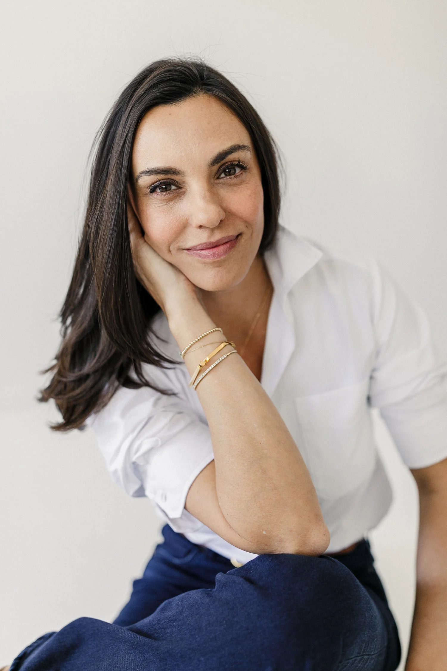 A woman with long dark hair, smiling, wearing a white button-up shirt and gold jewelry, sitting against a plain white background.