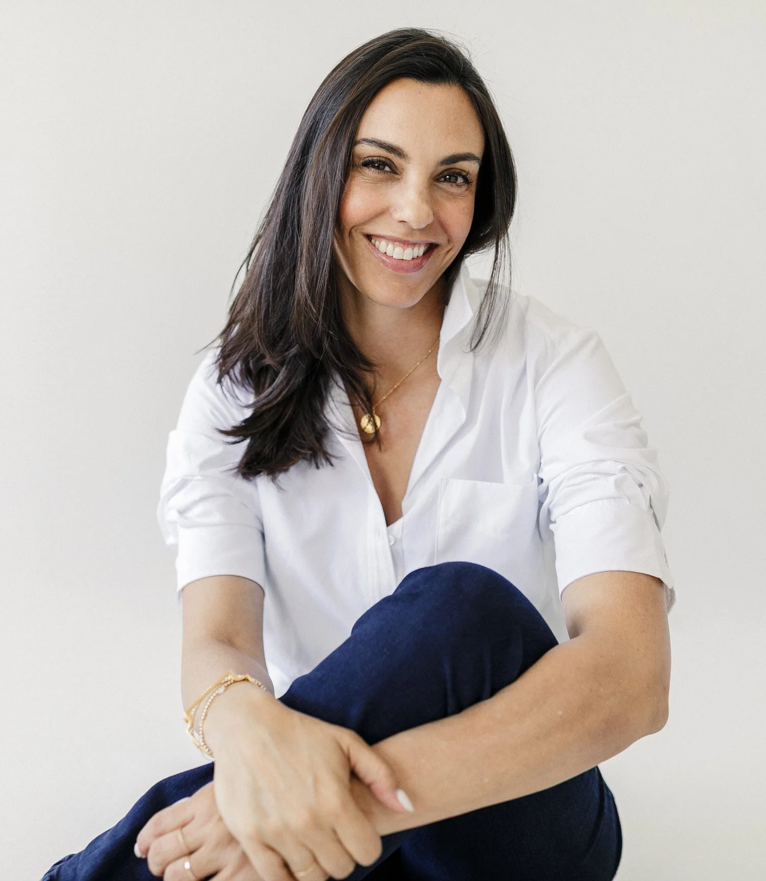 A woman with long dark hair, smiling and wearing a white shirt, sitting against a plain white background.