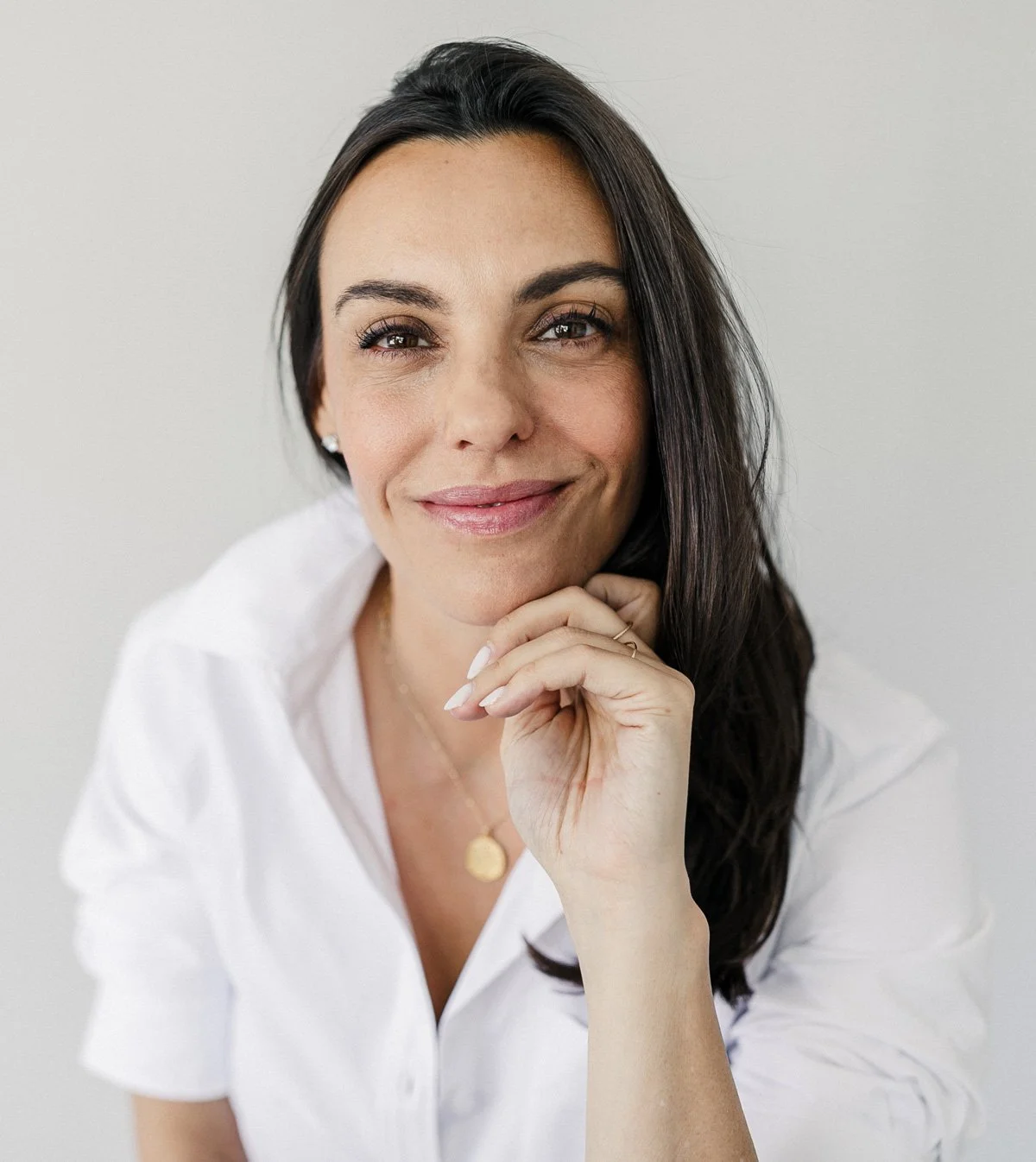 A woman with long dark hair, wearing a white top, smiling gently with her chin resting on her hand, appearing confident and friendly.