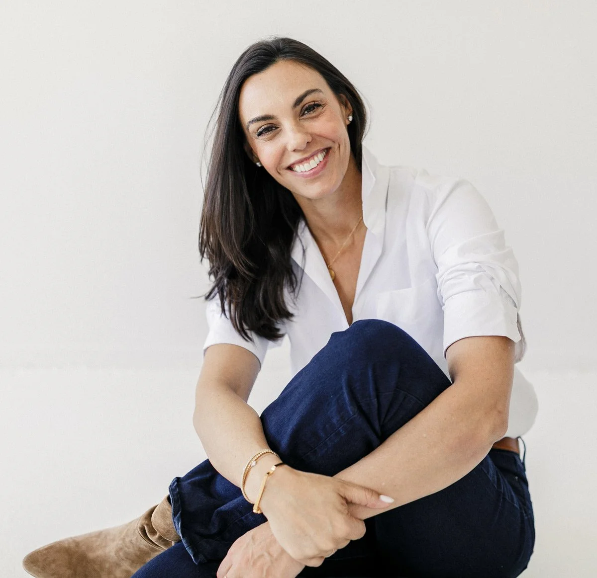 A smiling woman with dark hair, wearing a white collared shirt, dark blue jeans, and light brown boots, sitting against a plain light-colored wall.