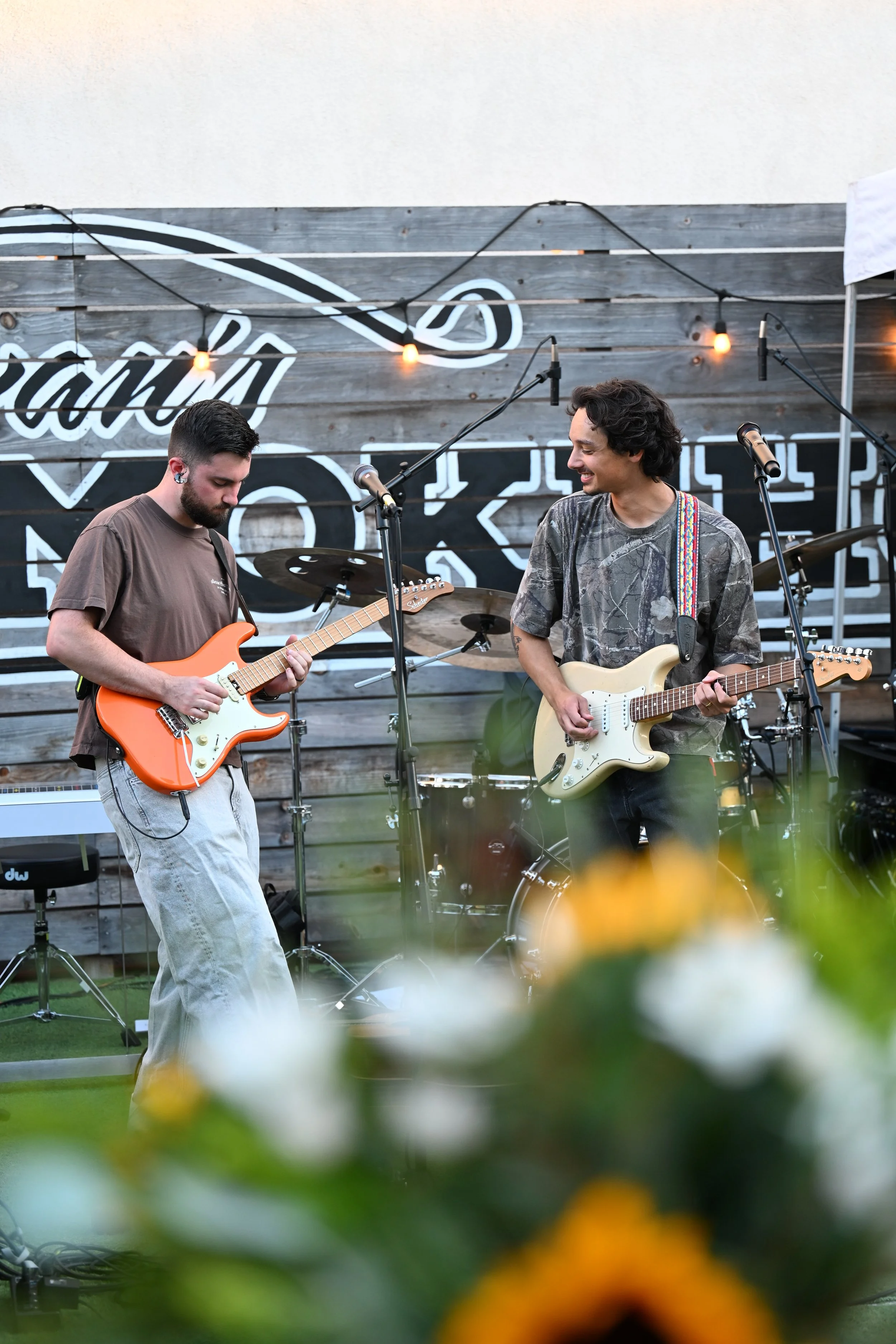 Two musicians playing electric guitars on an outdoor stage with a wooden backdrop and string lights. One is smiling at the other.