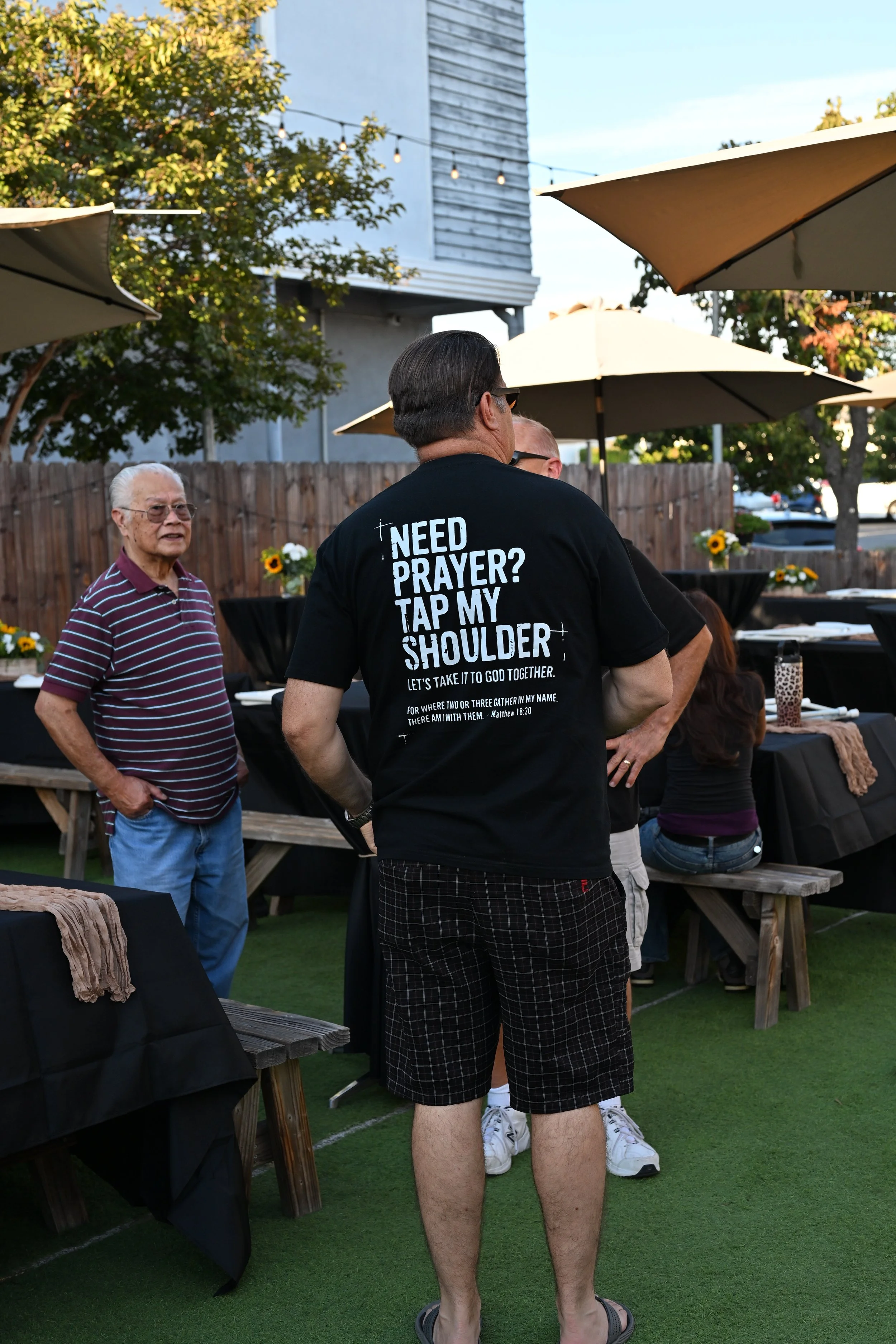 People gathered outdoors at a social event, standing around tables with black tablecloths and sunflower decorations, with umbrellas for shade, during daytime.