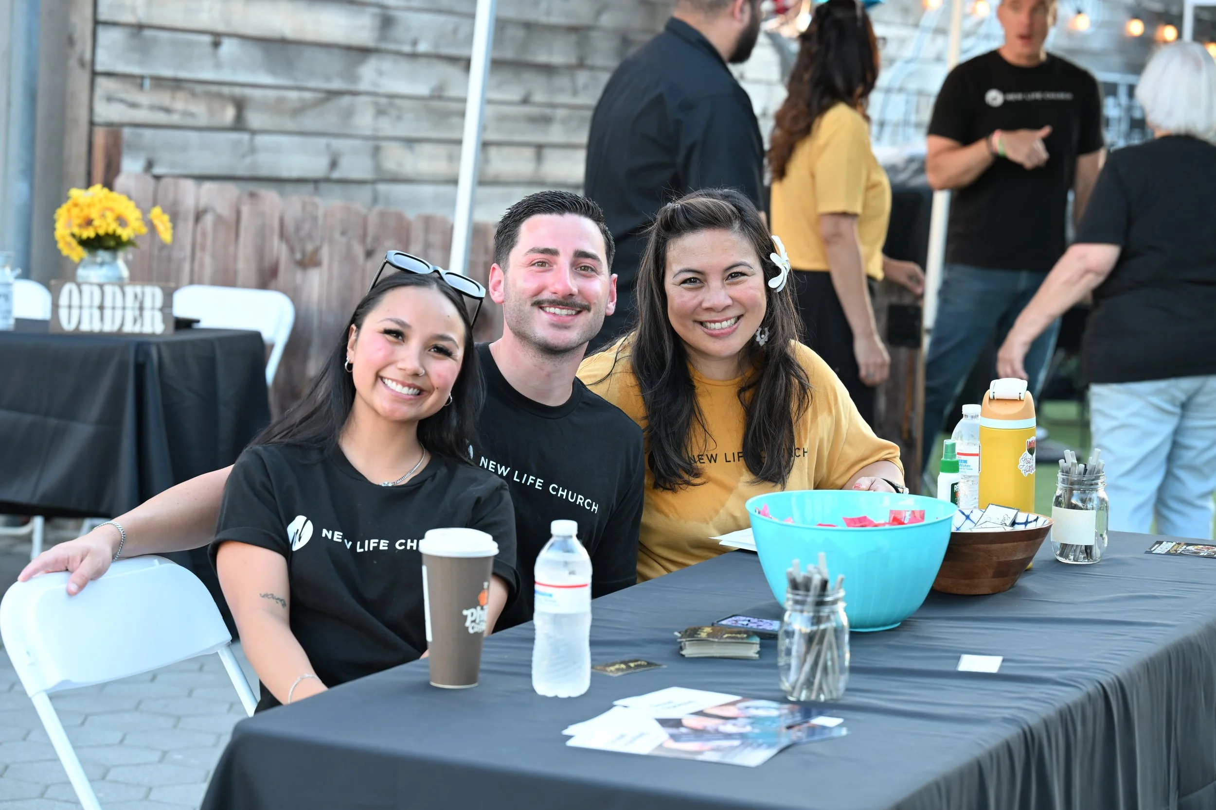 Three people sitting at a table outdoors, smiling and posing for the camera, with other people in the background at an event or gathering.
