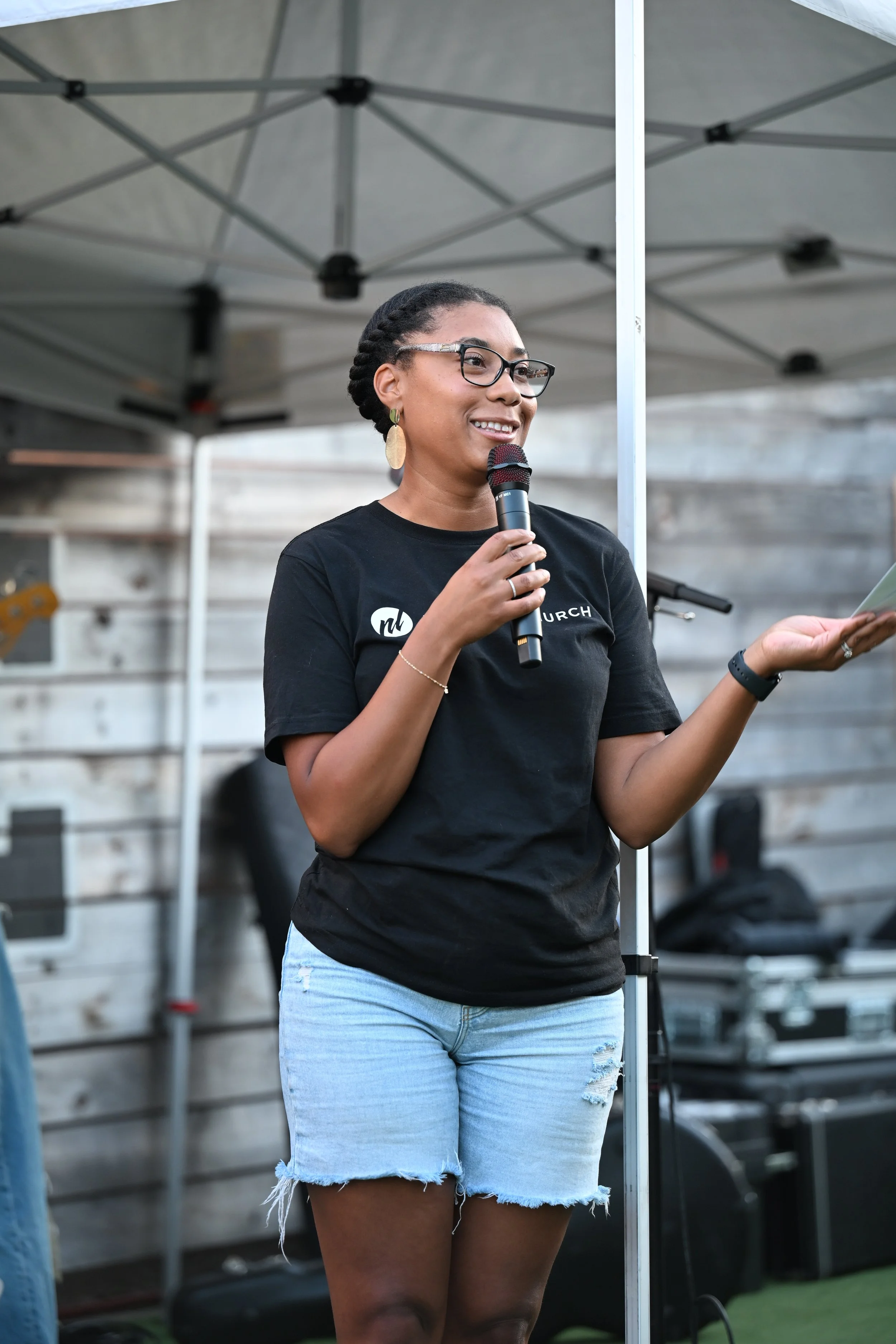 A woman with glasses, earrings, and a black T-shirt holding a microphone and a piece of paper, standing outdoors under a canopy.