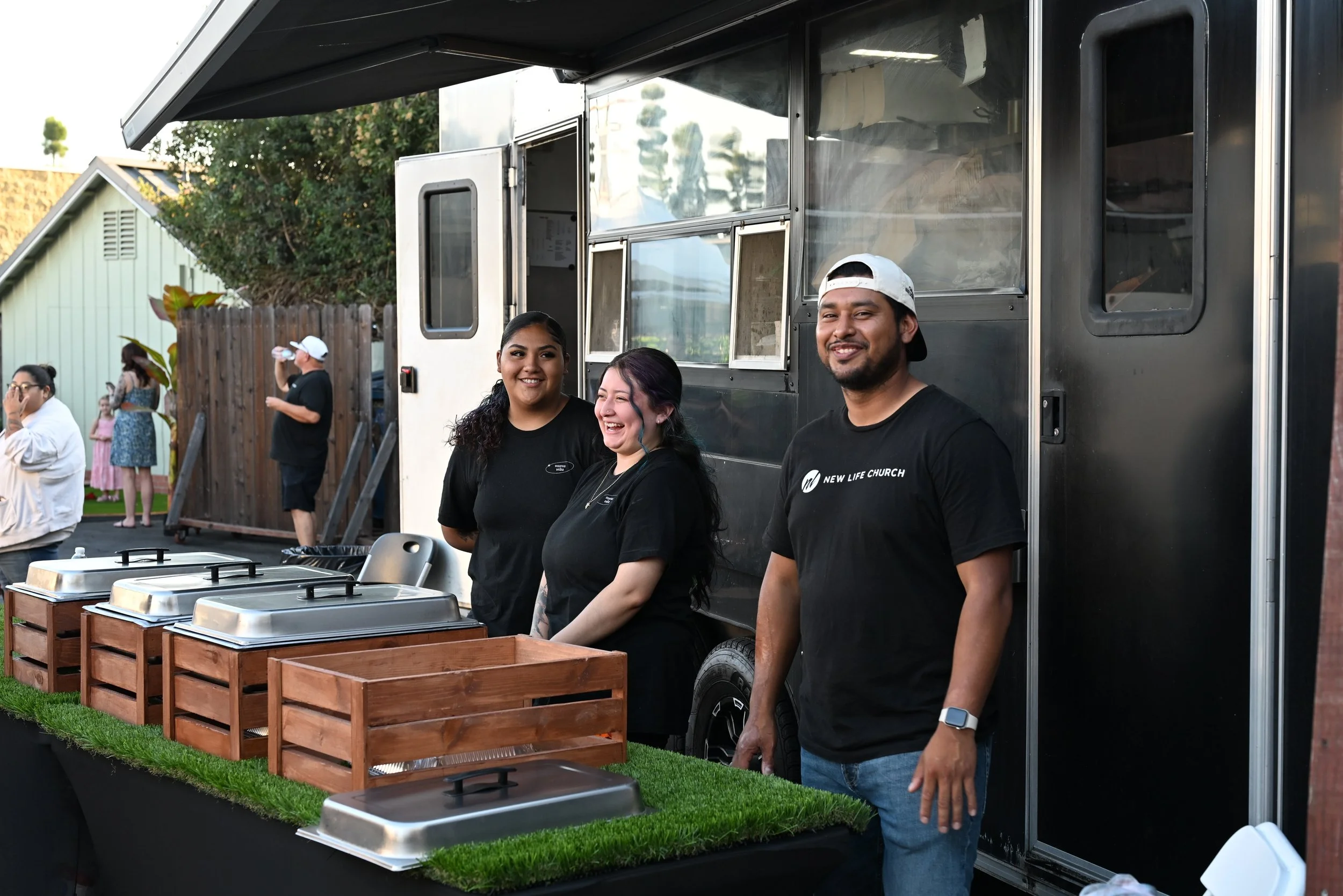 Three people stand in front of a food truck, smiling. The food truck has a black exterior with two open windows and a small serving counter. Two of them are women, and one is a man wearing a backwards cap, all wearing black shirts. In the background,