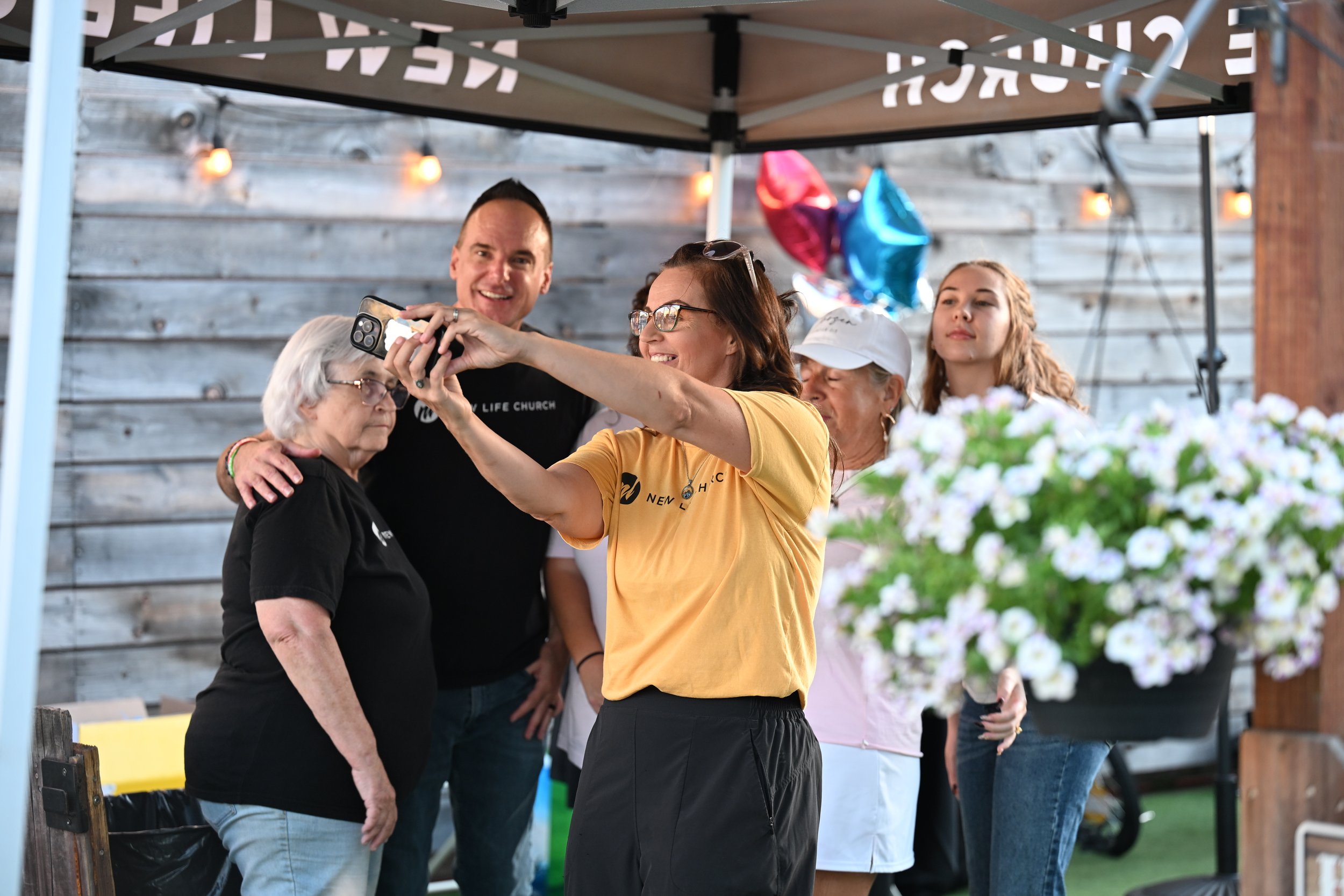 Group of people taking a selfie at an outdoor event, under a canopy with string lights, with balloons and flowers in the background.