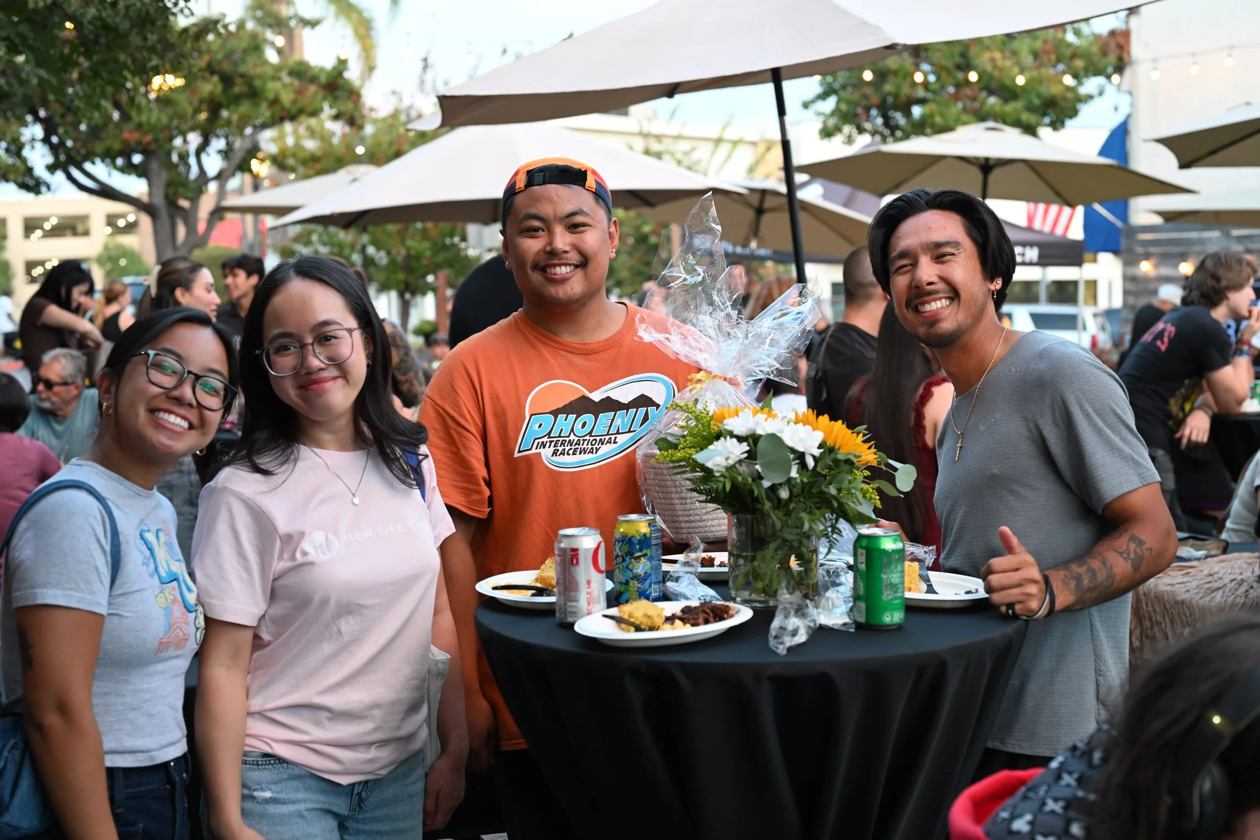 A group of four friends, two women and two men, smiling at an outdoor gathering. One woman and one man are wearing glasses. The man in the center is holding a bouquet of flowers and has a big smile. There are tables with food and drinks, and umbrella