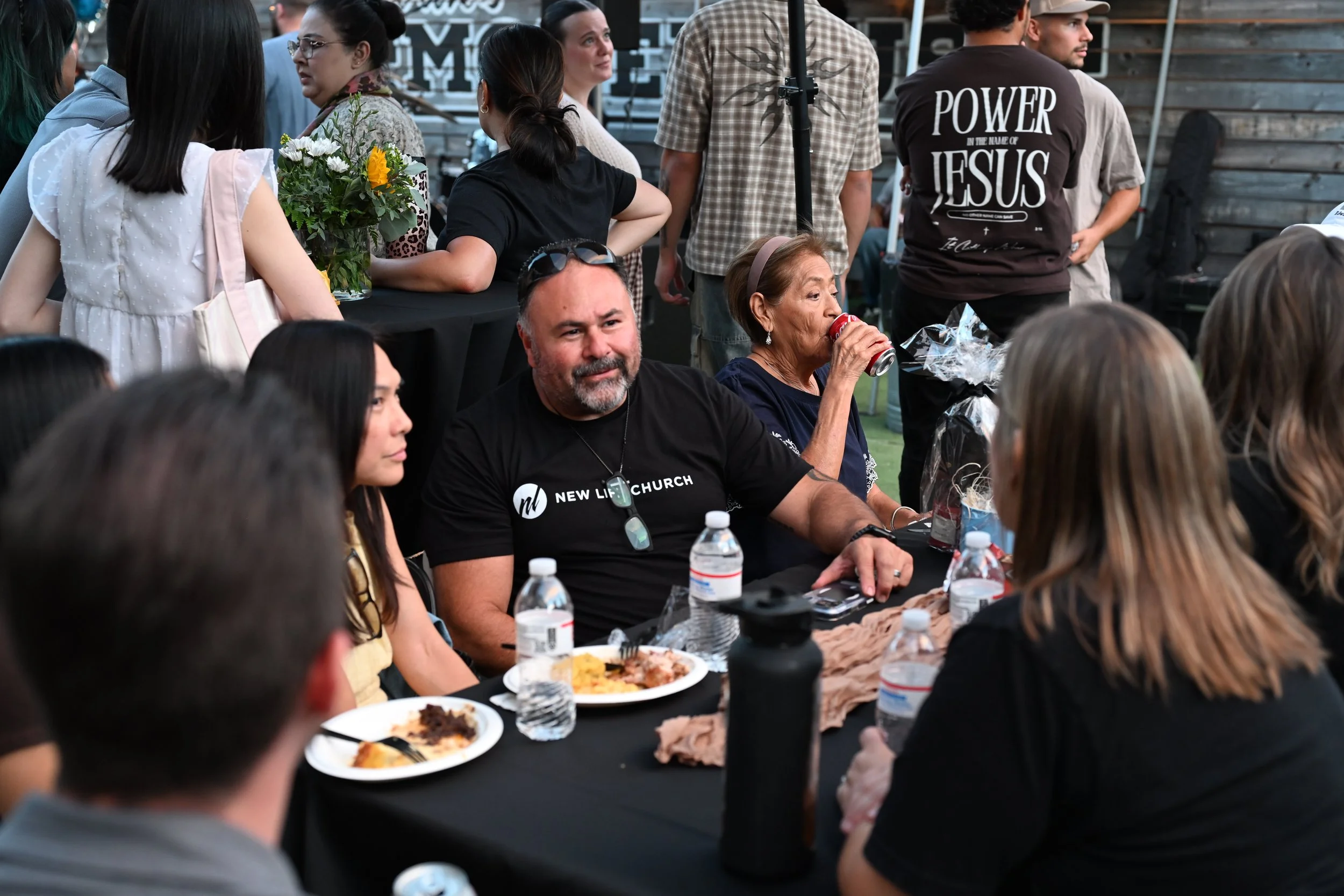 A group of people gathered around a table outdoors at a social event, with some eating and others talking. The table has plates of food, water bottles, and a bouquet of flowers. Several individuals are visible, including a woman drinking from a can, 