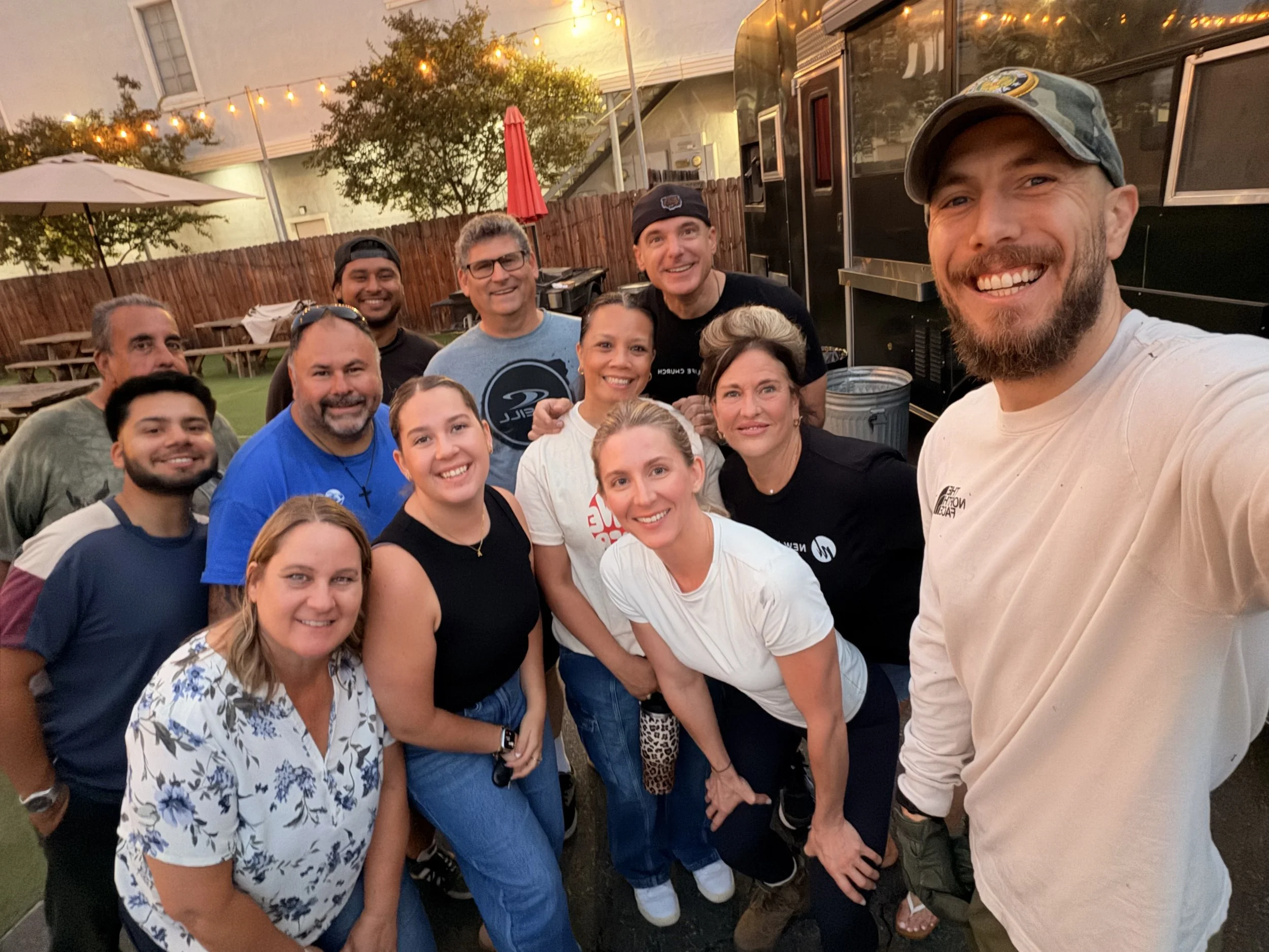 A group of people smiling and taking a selfie outdoors at night. There are string lights and trees in the background, along with a black food truck and picnic tables.