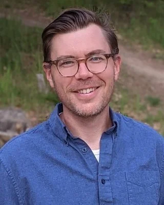 A smiling man with glasses and dark hair styled back, wearing a blue collared shirt, outdoors with grass and dirt in the background.