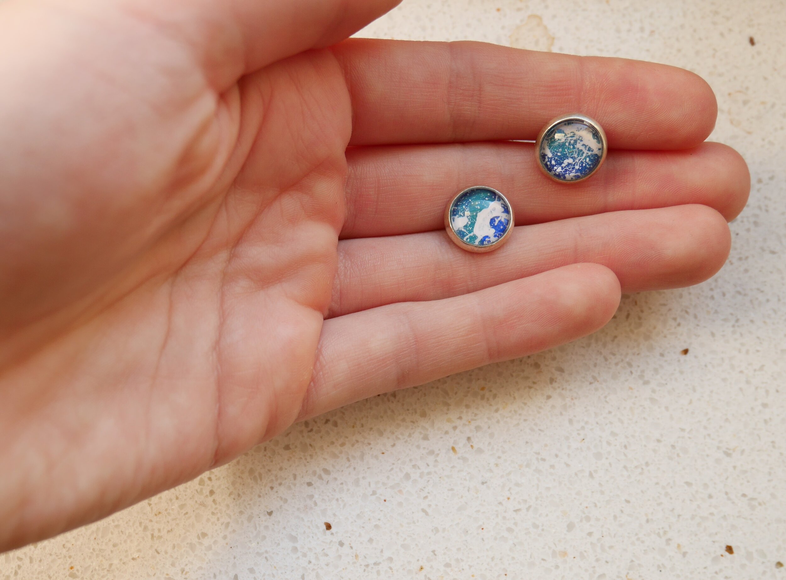 kleine, runde silberne Ohrstecker mit handbemalten Glascabochons in verschiedenen Blau- und Türkistönen und weißen Farbklecksen