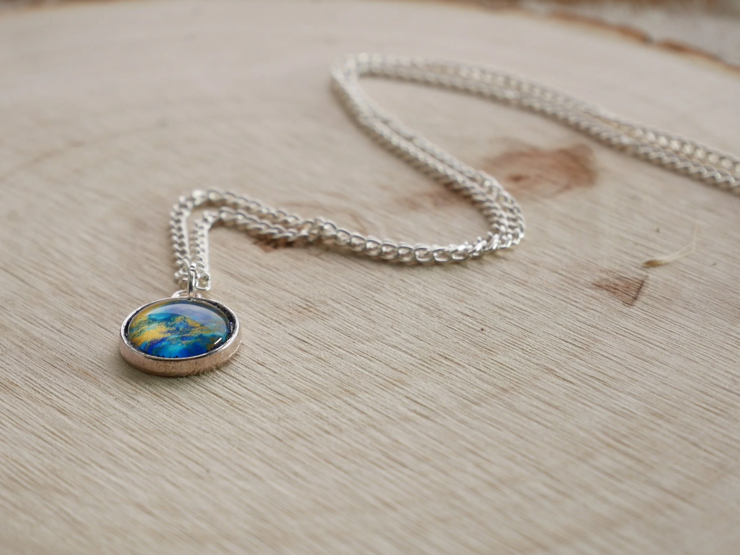 Silberne Halskette mit rundem Anhänger, darin eingefasst ein halbrunder Glasstein in blau, gold und glitzernd