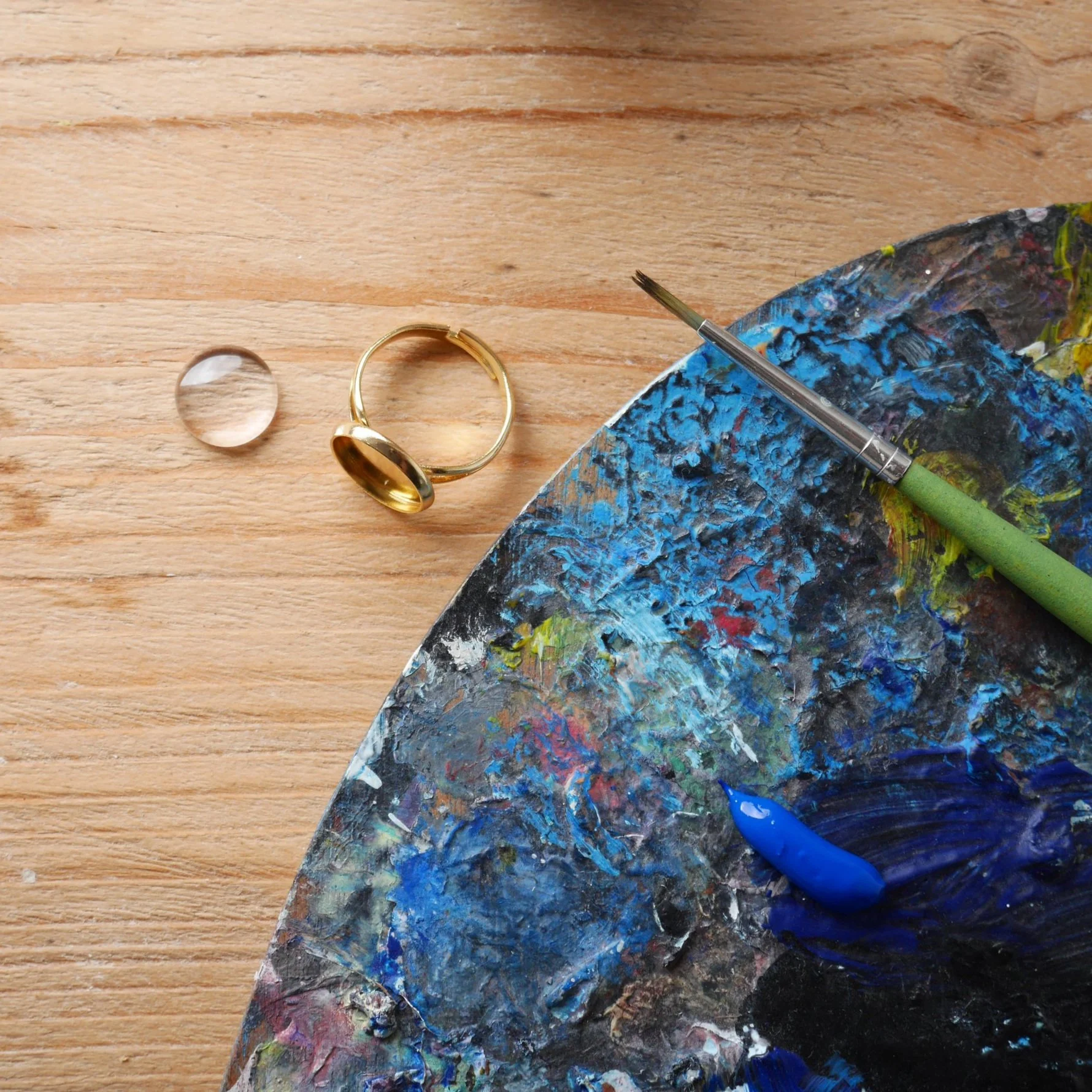 Kunstpinsel, blaue Farbe, Ring, Glasperle, Holzoberfläche.