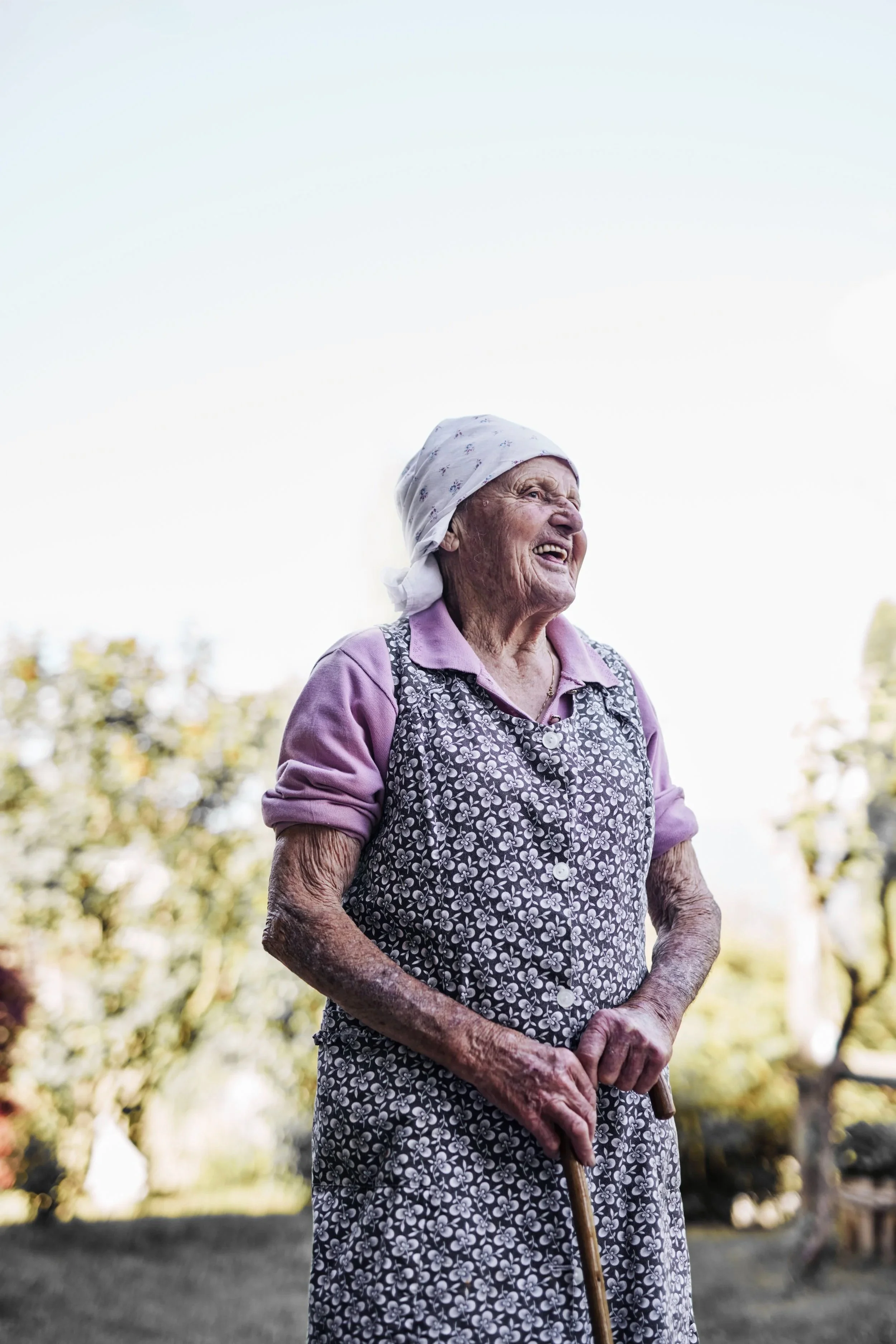 Elderly woman wearing a headscarf and floral dress, smiling outdoors.