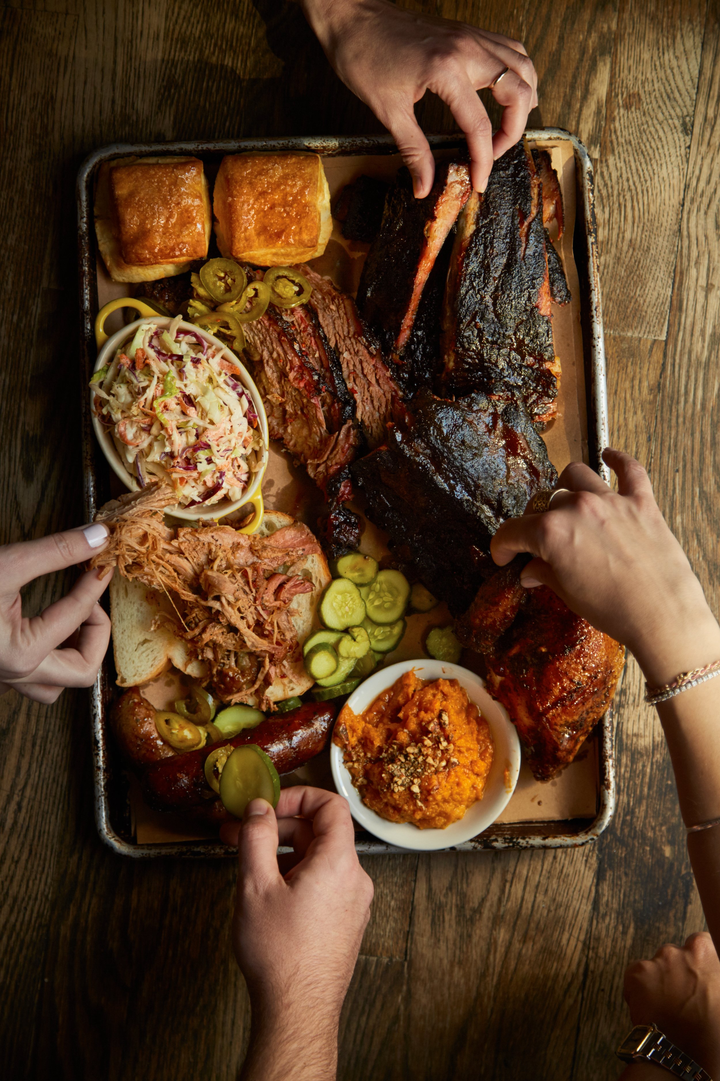 A wooden table with a tray of assorted barbecue meats including ribs, brisket, and sausage, accompanied by coleslaw, pickles, bread, sweet potatoes, and rolls. Multiple hands are reaching for the food.