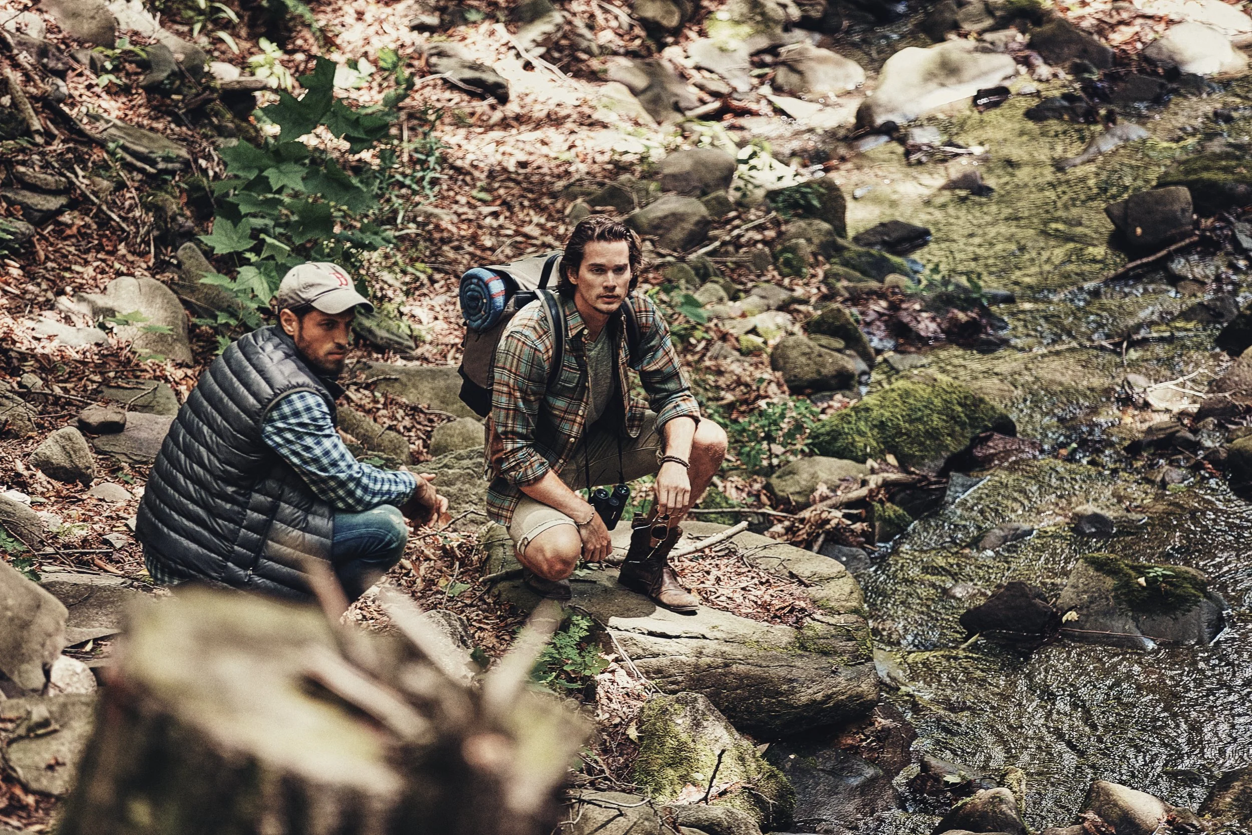 Two men sitting by a forest creek, wearing outdoor clothing, appearing to rest during a hike. One wears a hat and a vest, while the other has a backpack with a rolled-up sleeping pad.