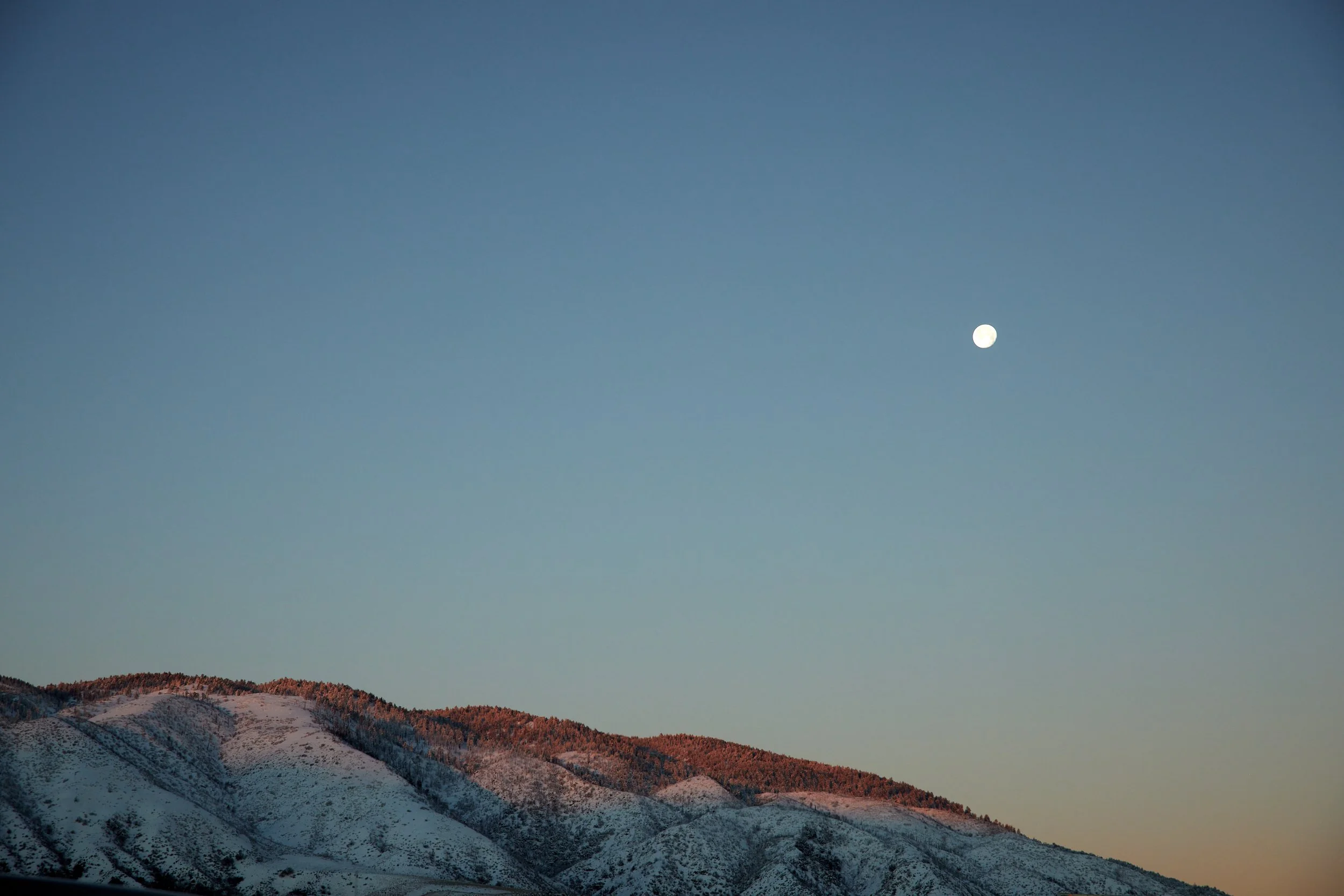 Snow-covered mountain with the moon in a clear sky at sunset, creating a serene natural landscape.