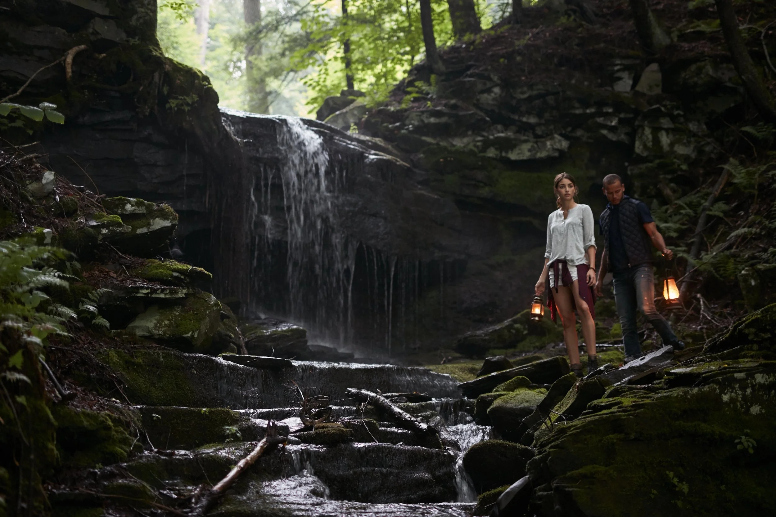 Two people holding lanterns in a forest by a waterfall, exploring a moss-covered rocky area.