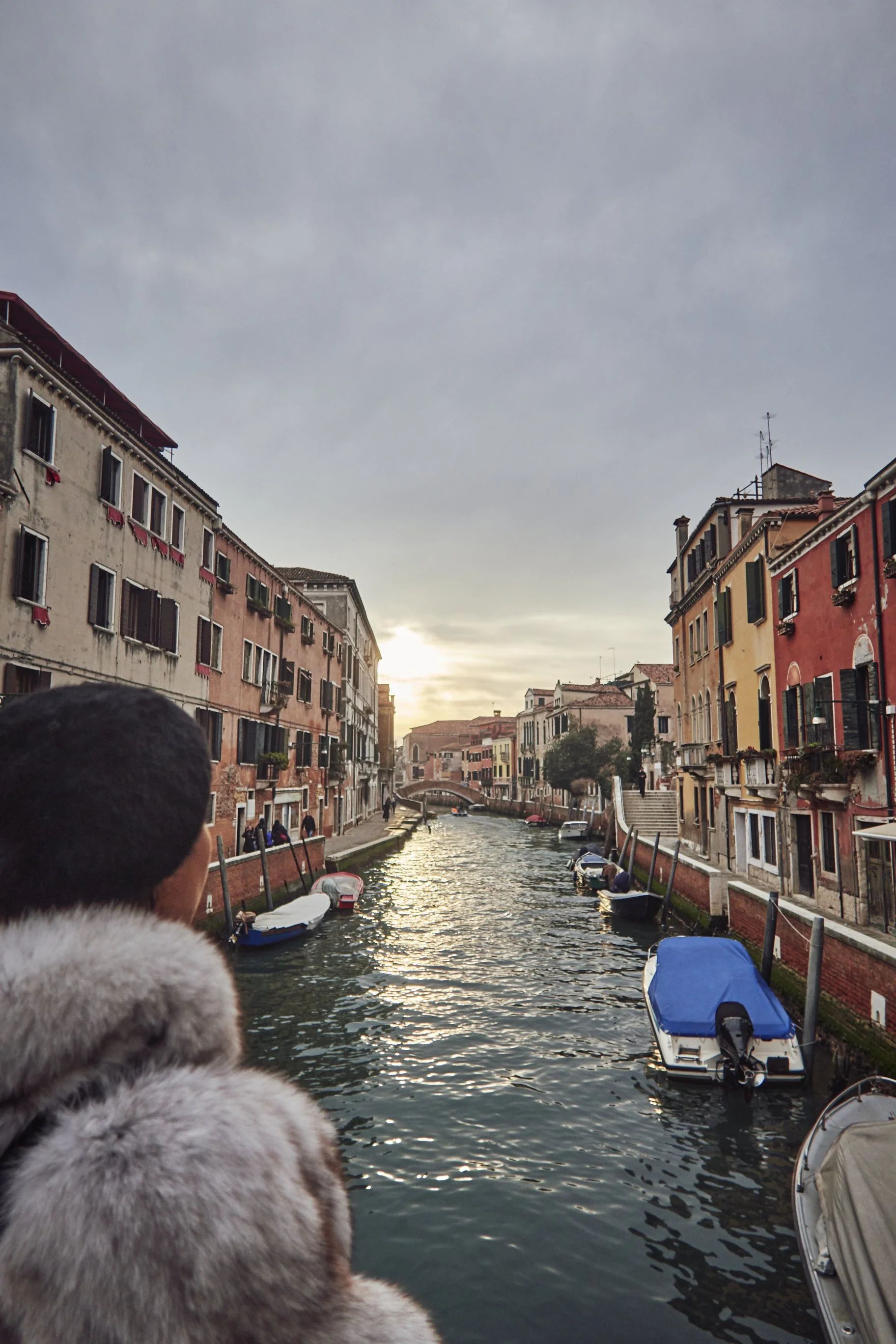View of a Venetian canal at sunset with boats and colorful buildings lining both sides, and a person wearing a fur-covered hood in the foreground.