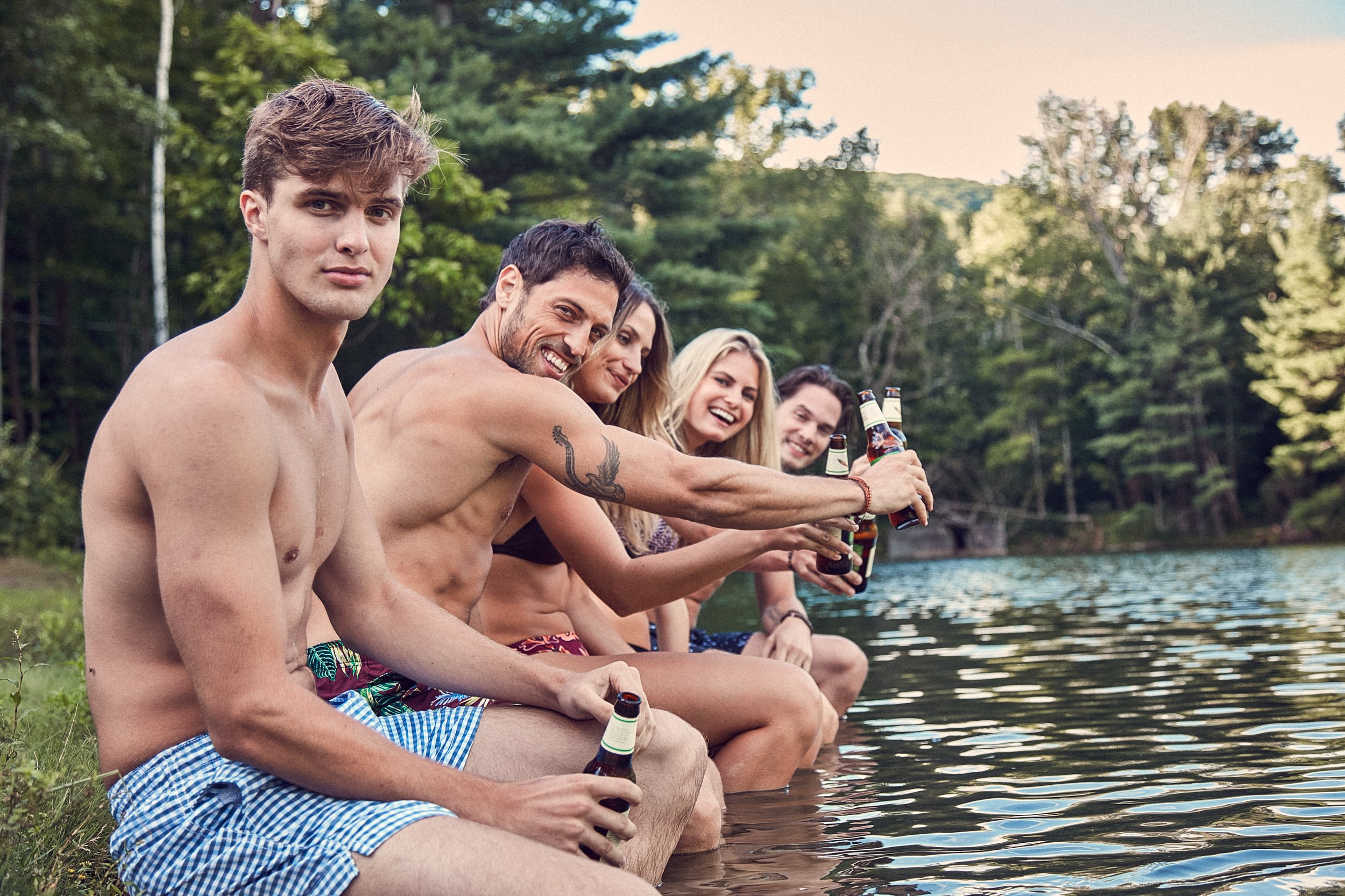 Group of young adults sitting by a lake, wearing swimsuits, holding bottles, and smiling with trees in the background.