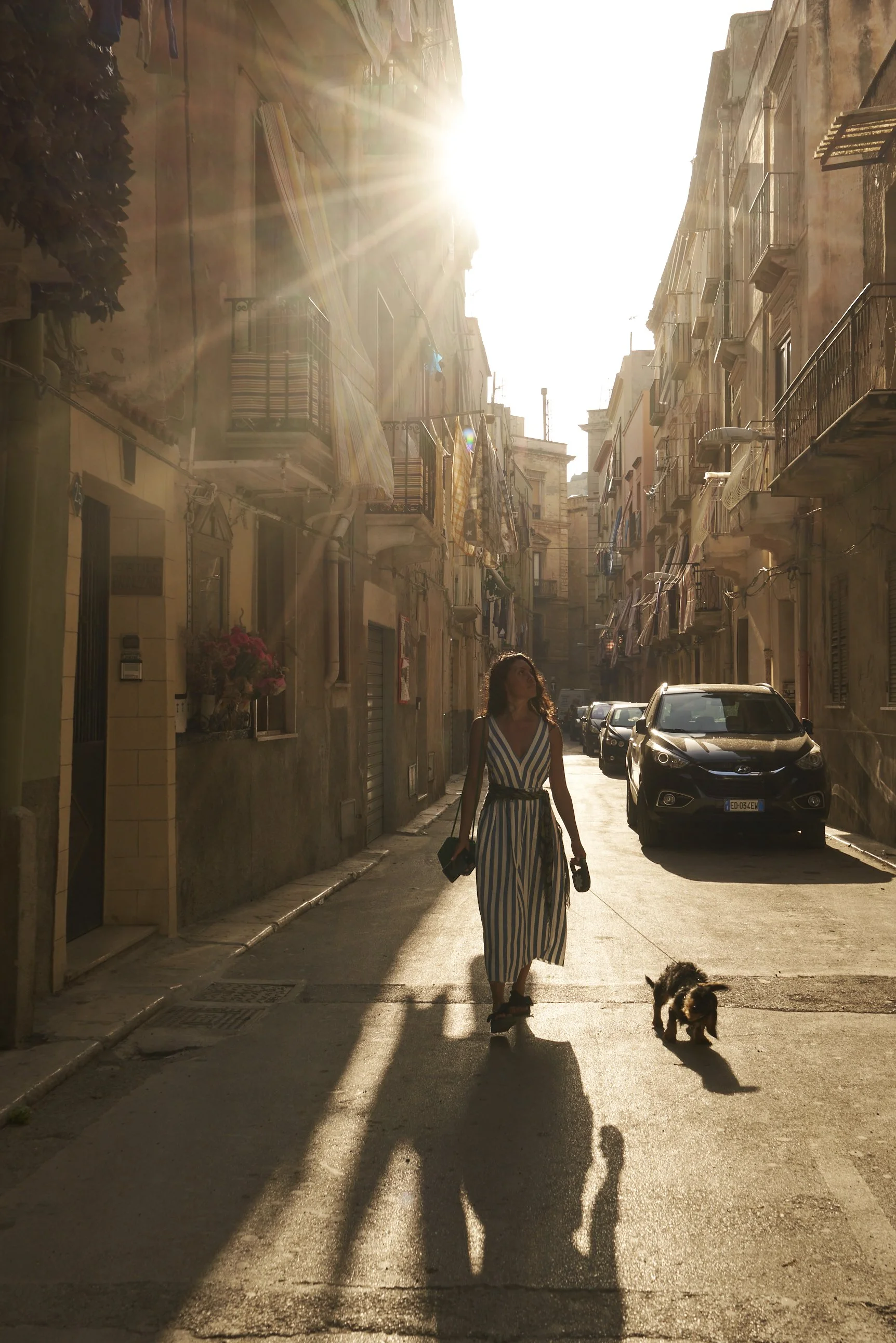 Woman walking a dog in a narrow sunlit street with old buildings and parked cars; sun rays creating dramatic shadows.