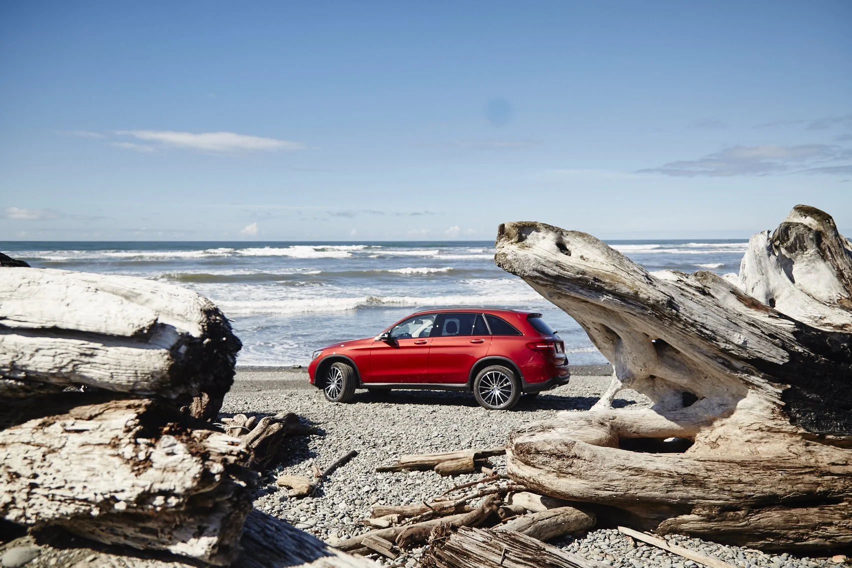Red SUV parked on a pebble beach near large driftwood, with ocean waves and blue sky in the background.
