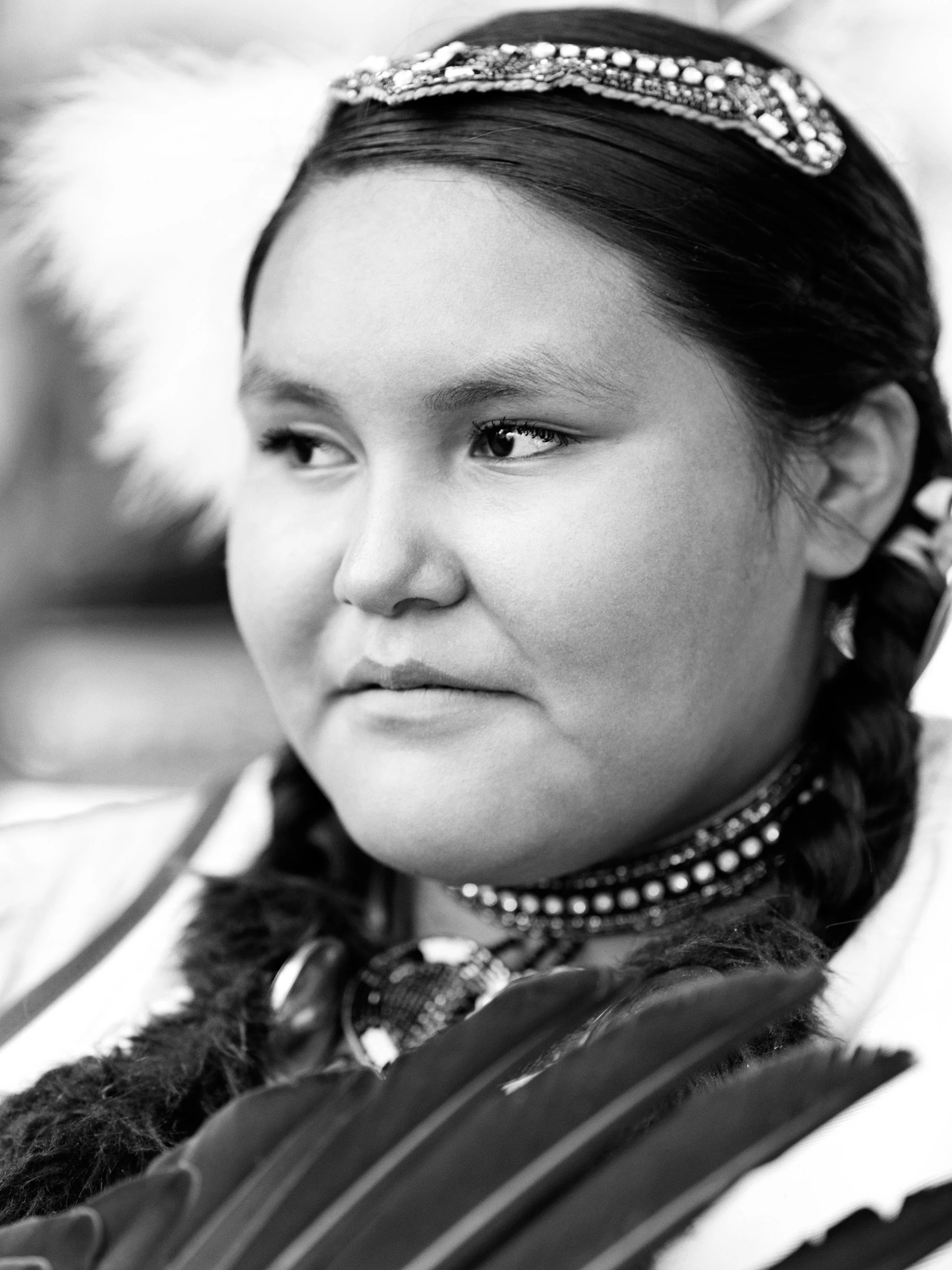Black and white portrait of a person wearing traditional clothing, including a beaded headband and feather decorations.