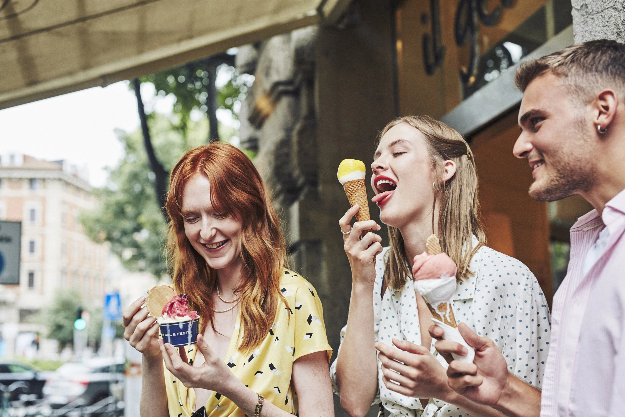 Three people enjoying gelato outdoors, two with cones and one with a cup, smiling and dressed in casual clothing.