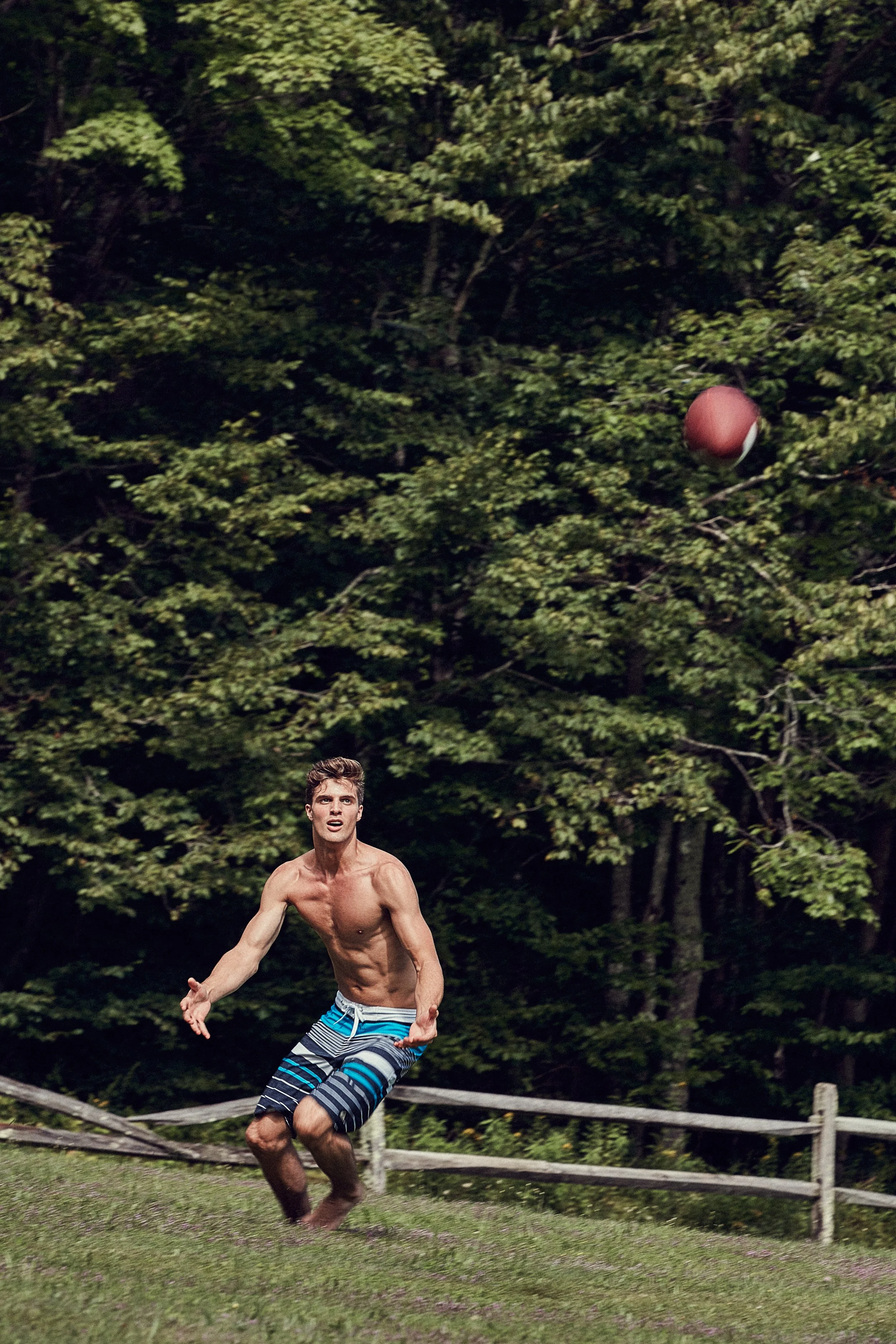 Shirtless man playing catch with an American football in a grassy field with trees in the background.