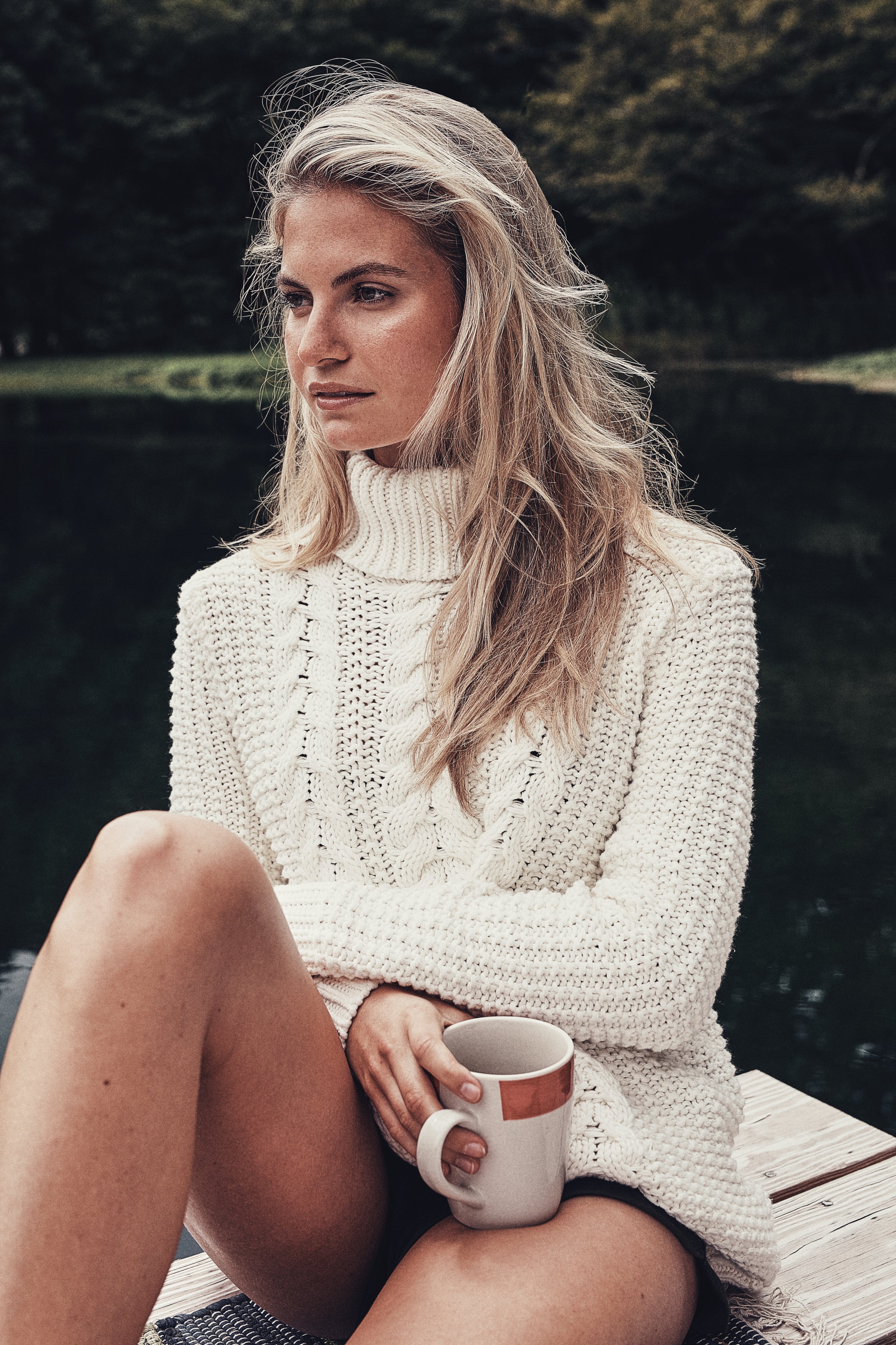 Woman wearing a white knitted sweater sitting near water, holding a mug.