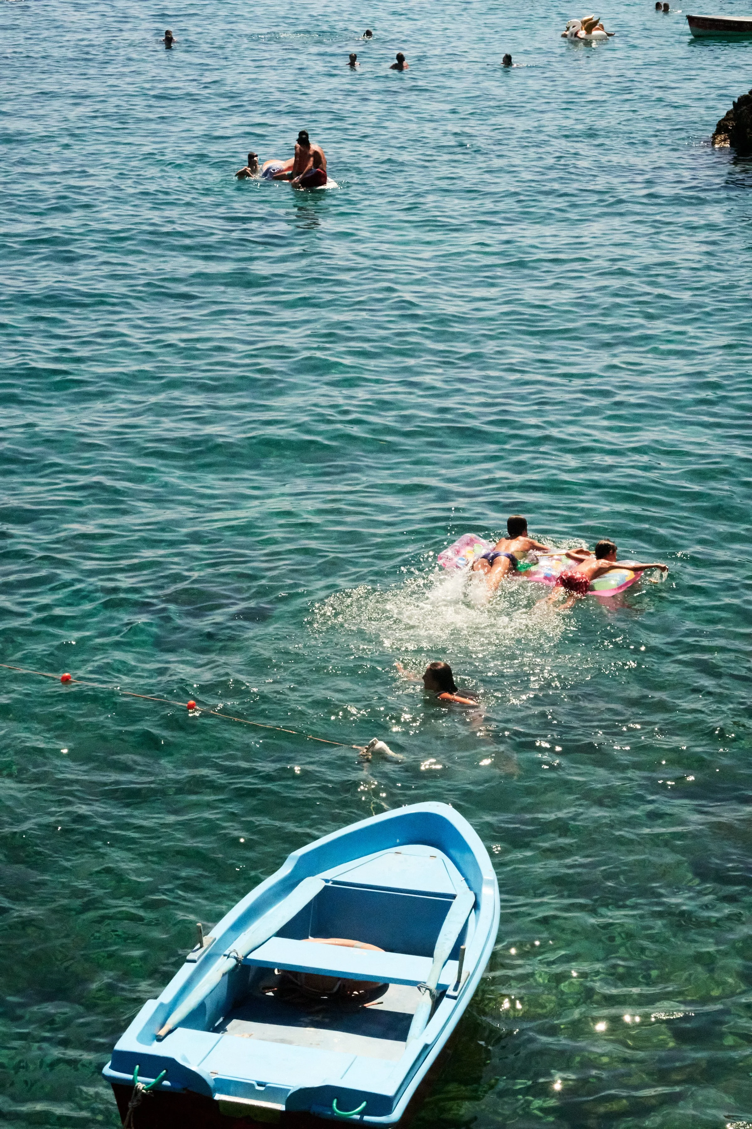 People swimming and playing on inflatable rafts in the ocean, with a small blue rowboat floating nearby.