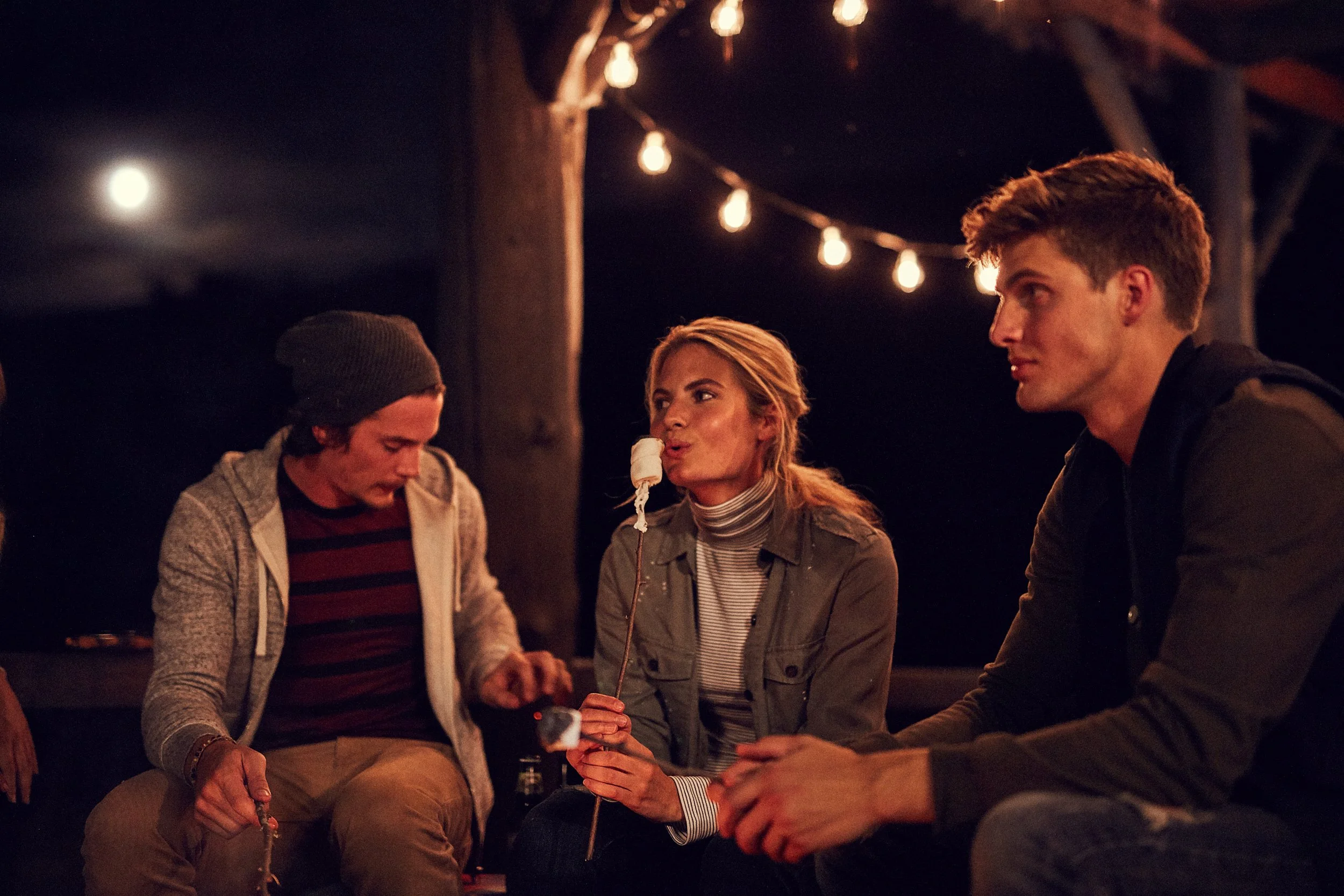 Three people sitting around a campfire at night, roasting marshmallows on sticks, with string lights and a full moon in the background.